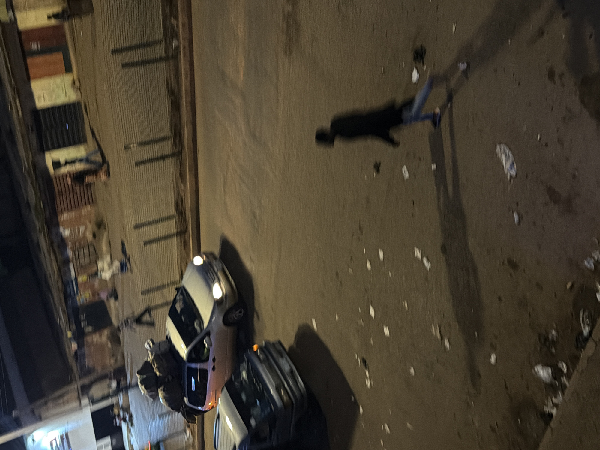 The image depicts a street scene at night in Bukuru, Nigeria, characterized by low light and motion blur, suggesting a dynamic environment. Two vehicles are visible on the asphalt road. The foreground vehicle, a dark colored van, is captured in motion, with its headlights illuminated. Behind it, a silver minivan is also in motion, its headlights on, and its roof laden with large, dark-colored bundles, possibly luggage or goods.
In the midground, several figures are present on a sidewalk or elevated area adjacent to the road. One figure, dressed in dark clothing, appears to be walking away from the camera. Further back, other indistinct figures are visible, some seemingly standing or walking. The area behind the figures is lined with structures that appear to be market stalls or shops, with various colored panels on their facades and some dark entrances. A metal fence runs along the edge of the sidewalk.
The road surface is strewn with numerous pieces of litter, primarily white and light-colored paper or plastic debris. A solitary, silhouetted figure is prominently featured in the lower right portion of the image, walking away from the camera, casting a long shadow. This figure is wearing a dark top and light-colored trousers. The overall impression is one of nighttime activity in a commercial or transportation hub, with elements of movement and disarray due to the presence of litter.