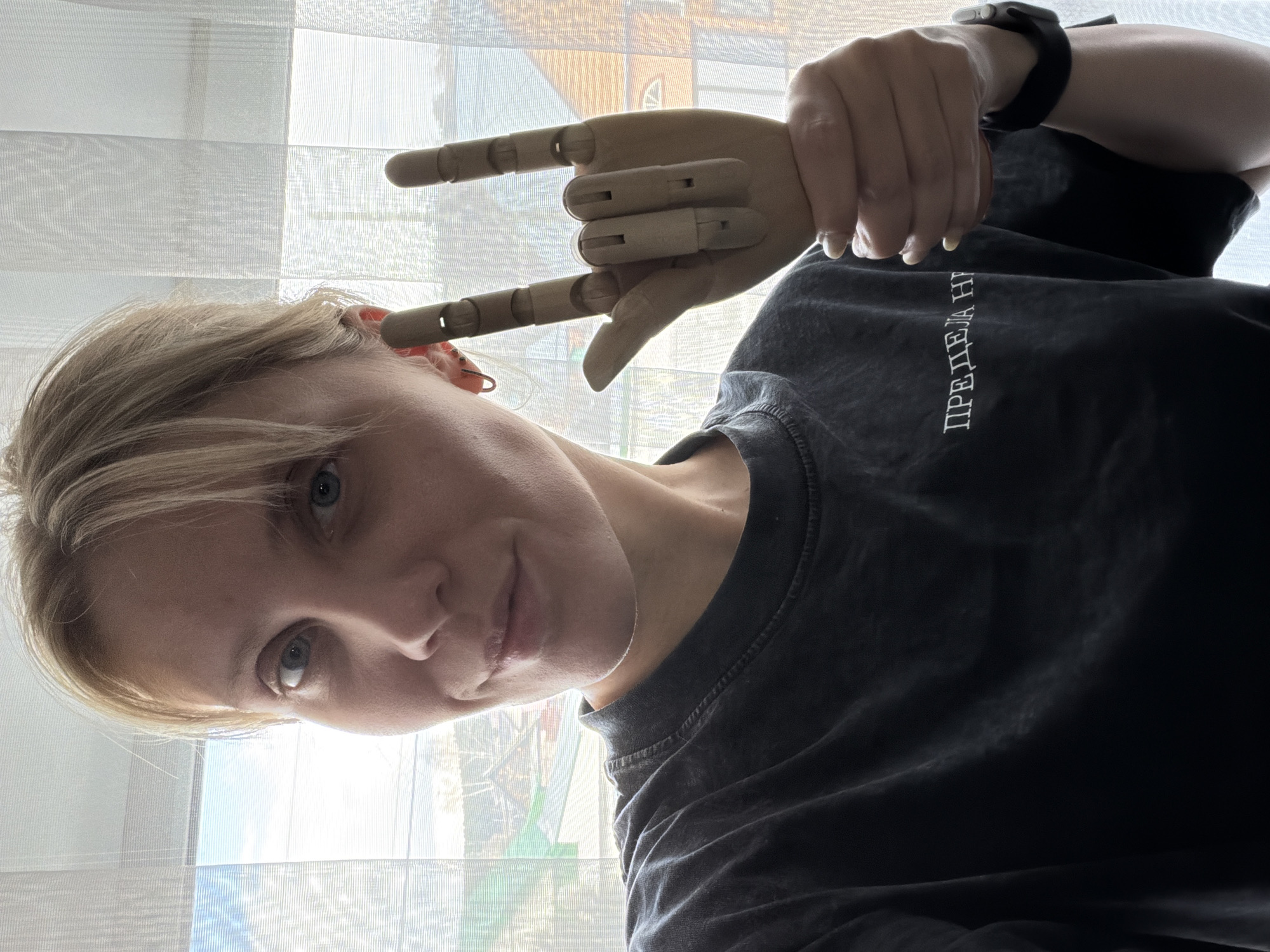 The image captures a vertical selfie of a person, likely a woman, with light blonde or light brown hair, looking directly at the camera. Her head is tilted towards the lower left of the frame, and her body extends upwards and to the right. She has a calm expression with a slight hint of a smile. The photo was taken indoors, positioned in front of a window covered with sheer white blinds or curtains, through which bright daylight is visible, suggesting it's daytime. Faint outlines of outdoor structures can be seen through the translucent fabric.
In the upper right quadrant of the image, the person's right hand holds a wooden, articulated artist's hand model. The model is arranged to display a gesture with the index and pinky fingers extended, the middle and ring fingers bent, and the thumb also extended, commonly known as the "rock on" sign or the "I love you" sign in American Sign Language. The person is wearing a dark-colored (likely charcoal grey or black) t-shirt. White Cyrillic text is printed vertically on the front of the shirt, which reads "ПРЕДЕЛА НЕТ," translating to "No Limit." A dark-banded smartwatch is visible on her left wrist. The location context for this image is Chaikovsky, Russia.