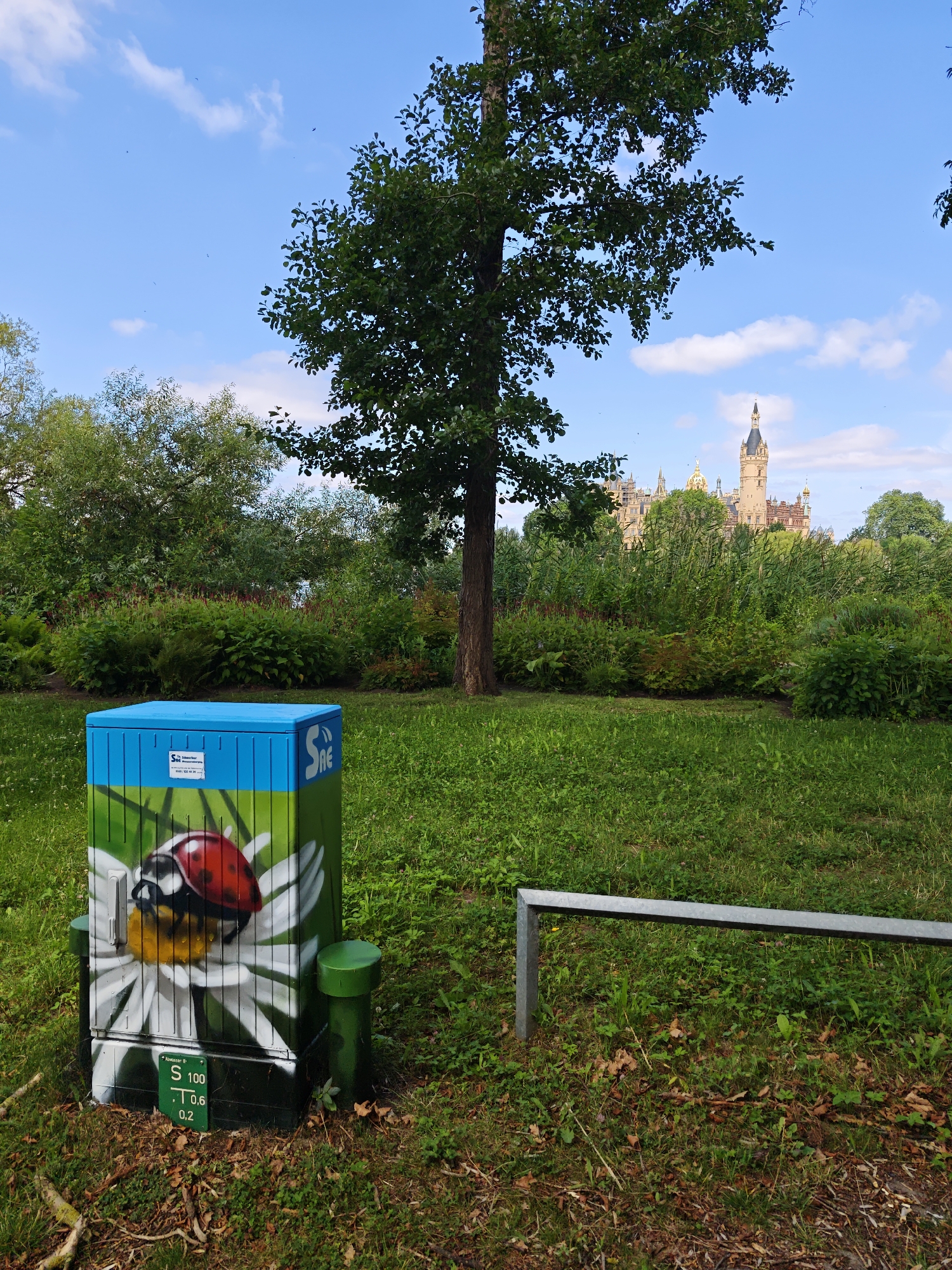 The image captures a vibrant green park with a blue and green utility box adorned with a painted ladybug and daisy in the foreground. Behind the box, a lush garden area with various plants and trees leads to a distant view of the Schwerin Castle, a prominent landmark in Schwerin, Germany. The sky is a clear blue with scattered white clouds. A metal railing is visible in the mid-ground to the right. The overall scene depicts a well-maintained outdoor space with a mix of natural elements and urban infrastructure.