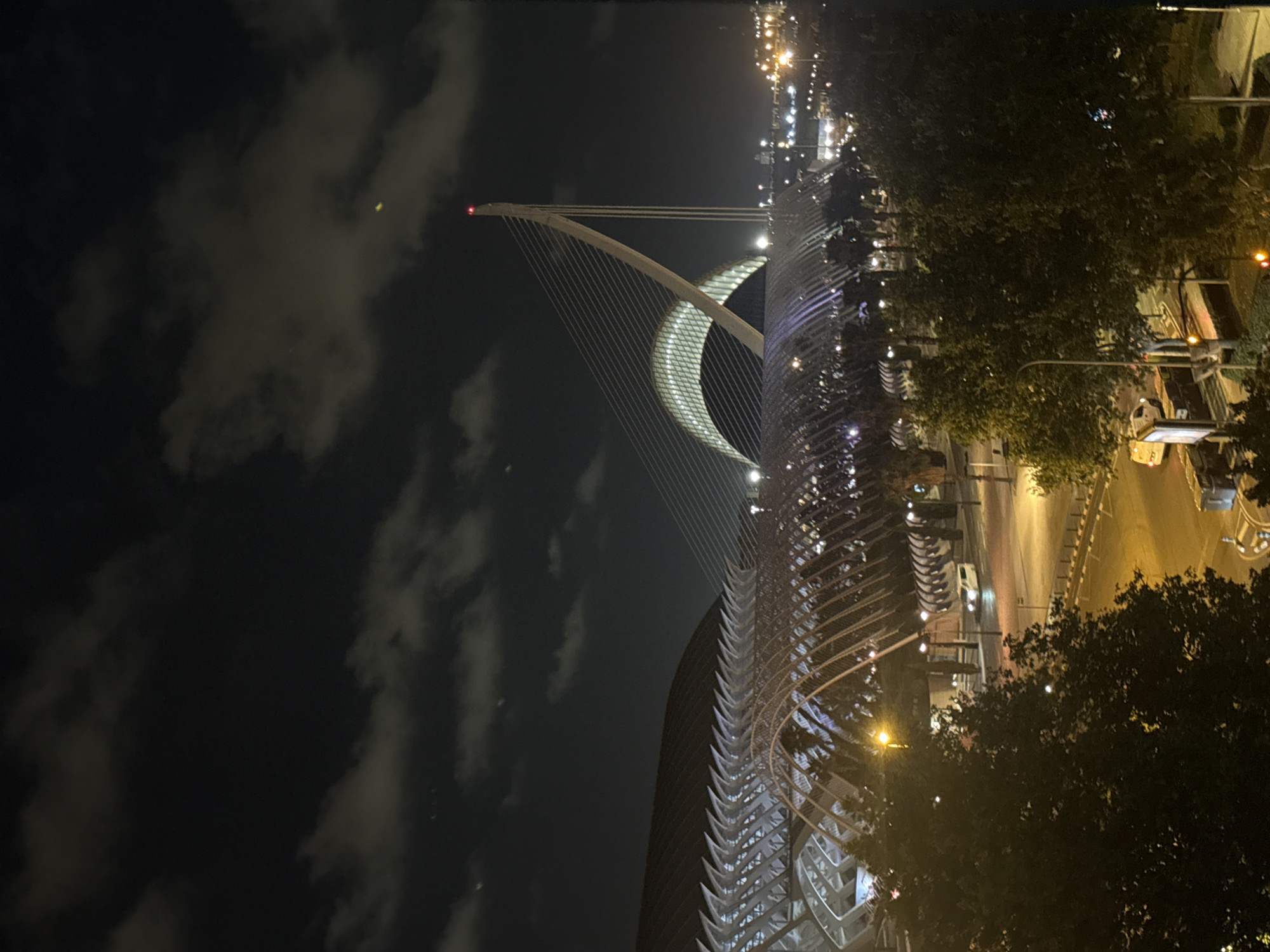 The image captures a nighttime view of the City of Arts and Sciences in Valencia, Spain. The architectural complex is illuminated, with the striking cable-stayed bridge dominating the scene. The bridge's white pylon, topped with a red light, stands tall against the dark, cloudy sky. The distinctive ribbed structure of the L'Hemisfèric is visible in the background, with its arched roof glowing with interior lights. In the foreground, dense trees with illuminated elements suggest parkland or pedestrian areas within the complex. A street with passing vehicles, including a white van, is visible below the structures, with streetlights casting a warm glow on the asphalt. The overall impression is one of modern, illuminated architecture set against a nocturnal urban landscape.
