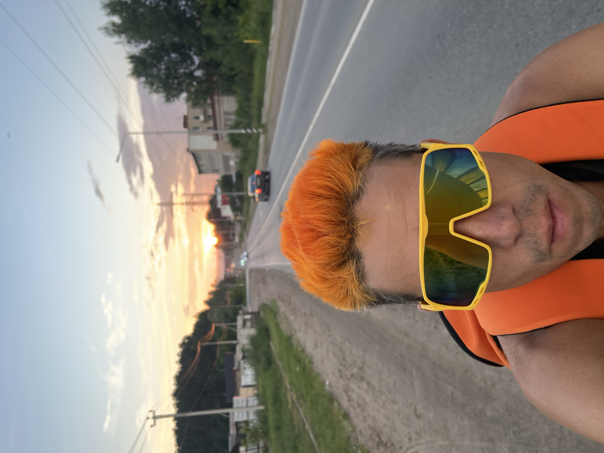 The image is a selfie taken from a low angle, featuring a man with bright orange hair and yellow sunglasses. He is wearing an orange vest or life jacket. The man is positioned in the foreground, with his head and shoulders dominating the frame.
In the background, a paved road stretches into the distance, bordered by a grassy shoulder and a dirt path on the left. Several cars are visible on the road, with their headlights on, suggesting it is either dusk or dawn. On the left side of the road, there are some low-rise buildings and a few scattered trees. On the right side of the road, more trees and some industrial-looking structures are present.
The sky is a soft gradient of blues, oranges, and pinks, indicating a sunset or sunrise. Power lines and poles are visible stretching across the landscape. The overall setting appears to be a rural or semi-urban area, possibly on the outskirts of the city of Chaikovsky, Russia, given the presence of industrial elements and the specific style of the low-rise buildings. The lighting suggests it is late evening or early morning.