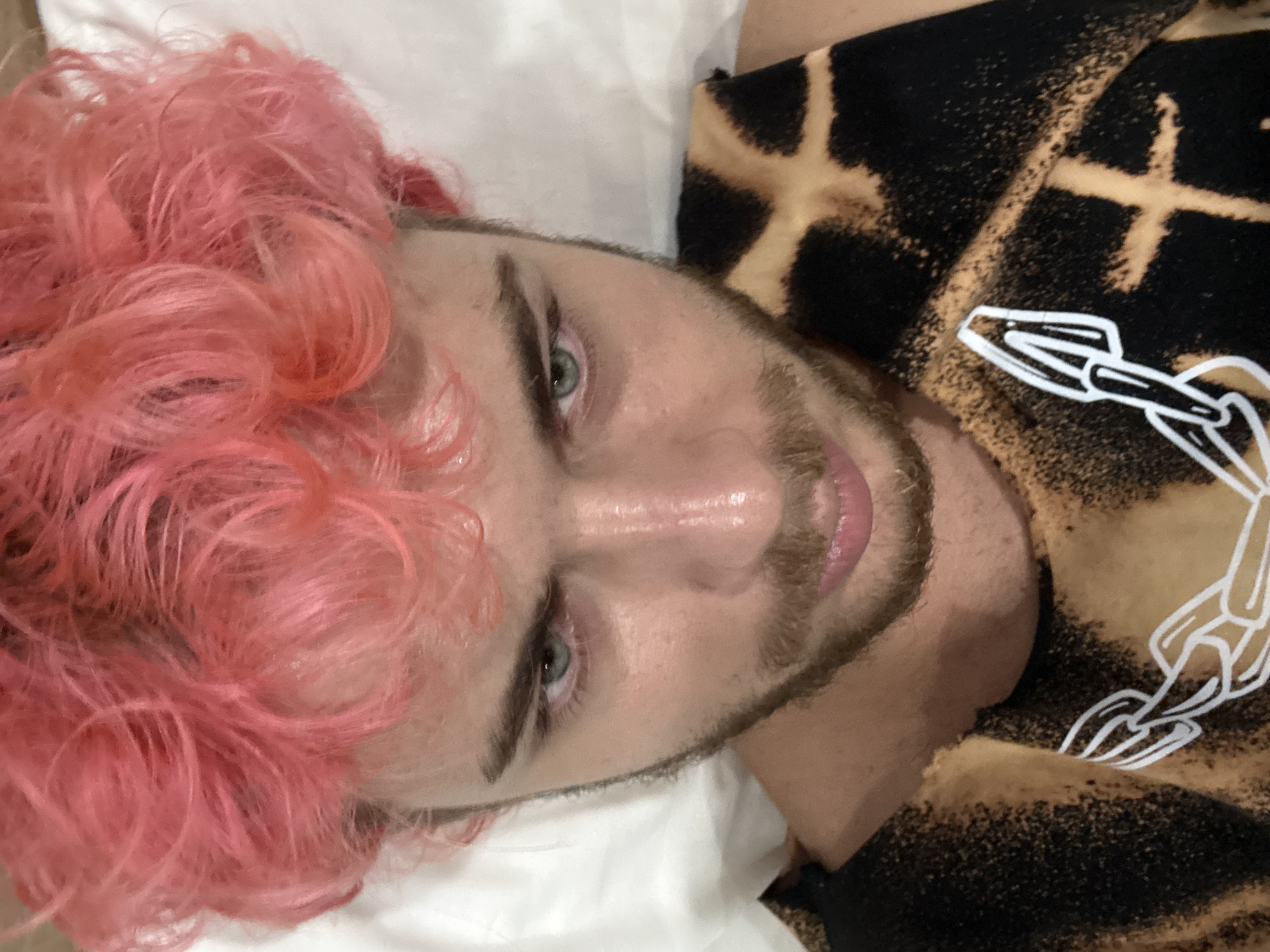A man with pink, curly hair is lying on a white pillow, looking at the camera. He has a beard and green eyes. He is wearing a black shirt with a bleach tie-dye pattern and a white graphic on the chest. The environment appears to be an indoor setting, possibly a bedroom. The city of Đà Nẵng, Vietnam, is not evident in the image.