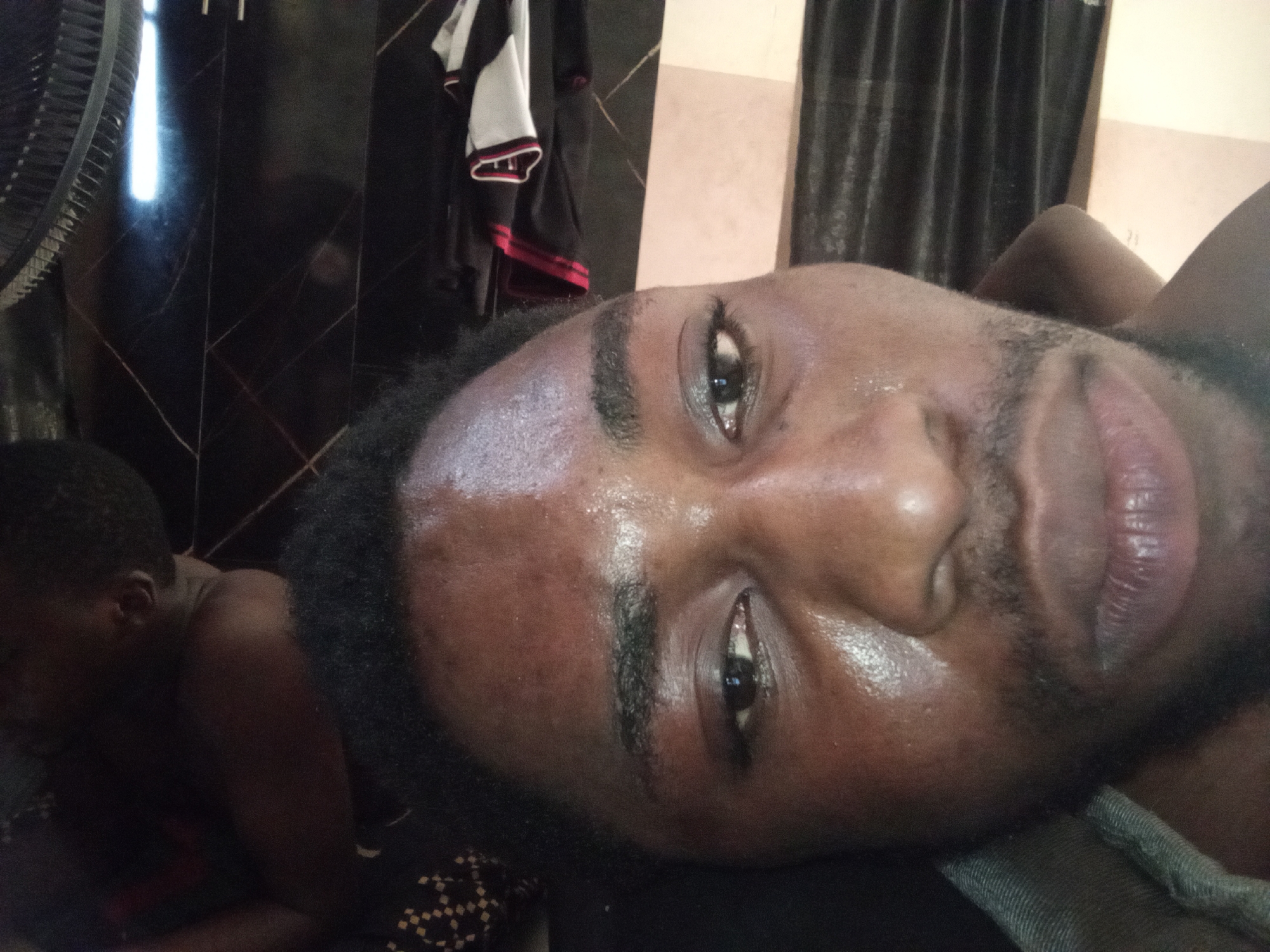 The image shows a close-up, low-angle shot of a young Black man's face, angled upwards. He is lying down, with his head tilted to the side and slightly towards the camera. His skin is dark and appears shiny with sweat. His eyes are open, and his mouth is slightly parted. He has dark, bushy eyebrows and a light beard. The background is dark and out of focus, with some indistinct objects, including what appears to be clothing hanging on a rack and dark curtains. To the left, another person's head and shoulder are visible, also in shadow. The overall lighting is dim, with a bright light source emanating from the upper left. The scene appears to be indoors.