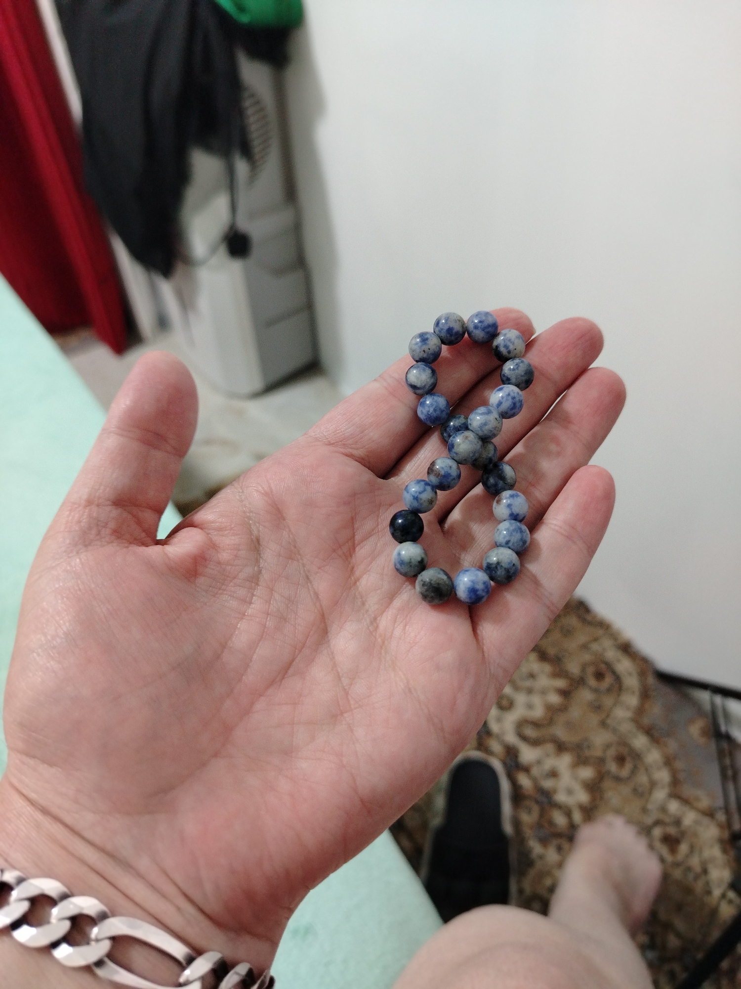 A person's left hand, palm up, holds a bracelet made of blue and white marbled beads arranged in a figure-eight shape. The hand is positioned in the foreground, with the wrist wearing a silver chain bracelet. In the background, out of focus, a light-colored wall and a patterned rug are visible, along with a black slipper and a bare foot. The overall lighting suggests an indoor setting. The city of Al Mas'oudi, United Arab Emirates, is not depicted in this image.