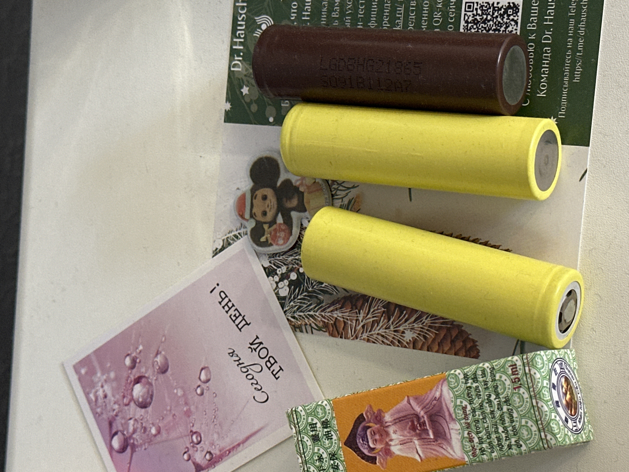The image shows a collection of objects arranged on a white surface, likely a desk or table. In the foreground, from left to right, is a small orange and green box with Asian script and a depiction of a seated figure in traditional garb. To its right are two cylindrical yellow objects, possibly batteries, positioned vertically. A third, dark brown cylindrical object, also possibly a battery, is placed to the right of the yellow ones.
Behind these objects, several pieces of paper or cards are visible. A pink card with water droplets and text in Russian, reading "Сегодня твой день!" (Today is your day!), is in the upper left. To its right is a small sticker of a cartoon character wearing a Santa hat and holding a gift. A green card with white text, partially obscured, is in the upper right, displaying "Dr. Hausch" and what appears to be a logo.
Further to the right, more text is visible on another green card, including "ka.ru/", and a QR code. The overall impression is of a still life arrangement of personal items, possibly documenting a collection or a moment. There is no indication of the city of Nevinnomyssk, Russia, within the image itself.