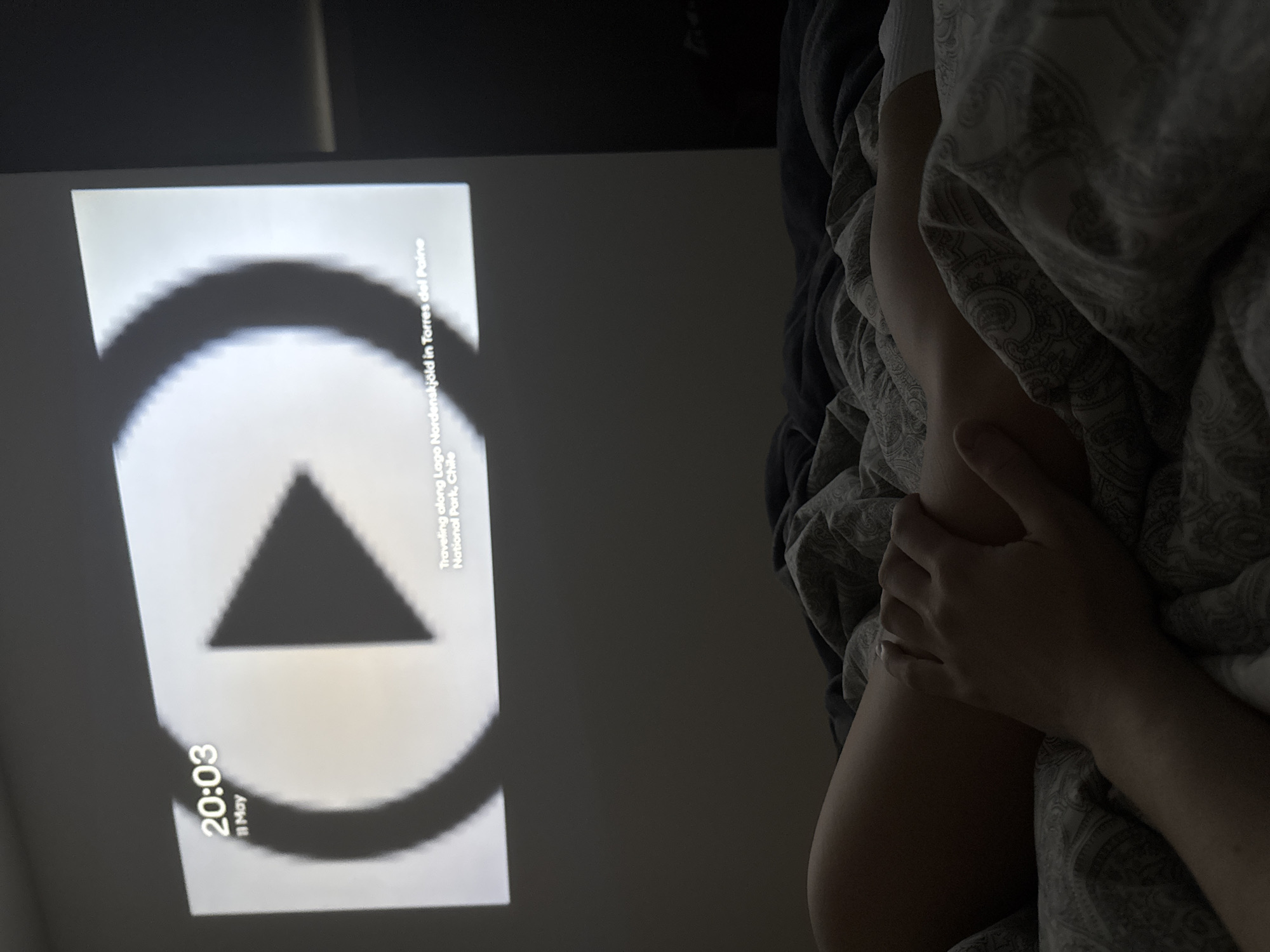 A person is in bed, covered with a duvet patterned with paisleys and other shapes. The person's hands are visible, and they appear to be holding the duvet. A video is playing on a screen, showing a triangular play symbol, overlaid with the time "20:03" and the date "11 May", and the text "Traveling along Lago Nordenskjöld in Torres del Paine National Park, Chile". The location is unspecified.