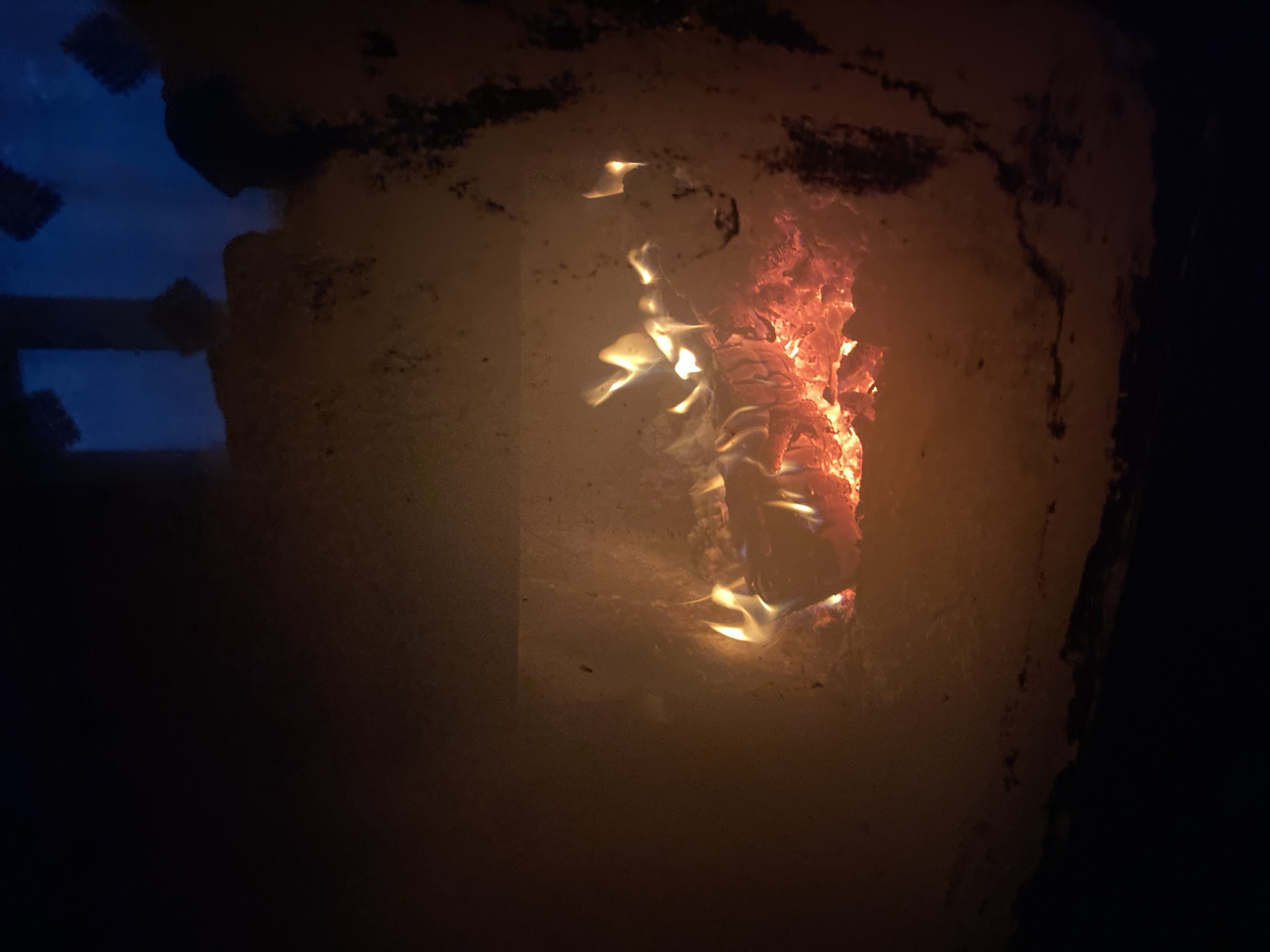 A view through the dirty glass door of a wood-burning stove reveals flames consuming pieces of wood. Outside, the dark outline of a window frame is visible in the upper left of the image, with some blue light suggesting it is night. No people are visible, and there is no indication of a specific location in Chaikovsky, Russia. The environment appears dark, with the stove being the only visible source of light.