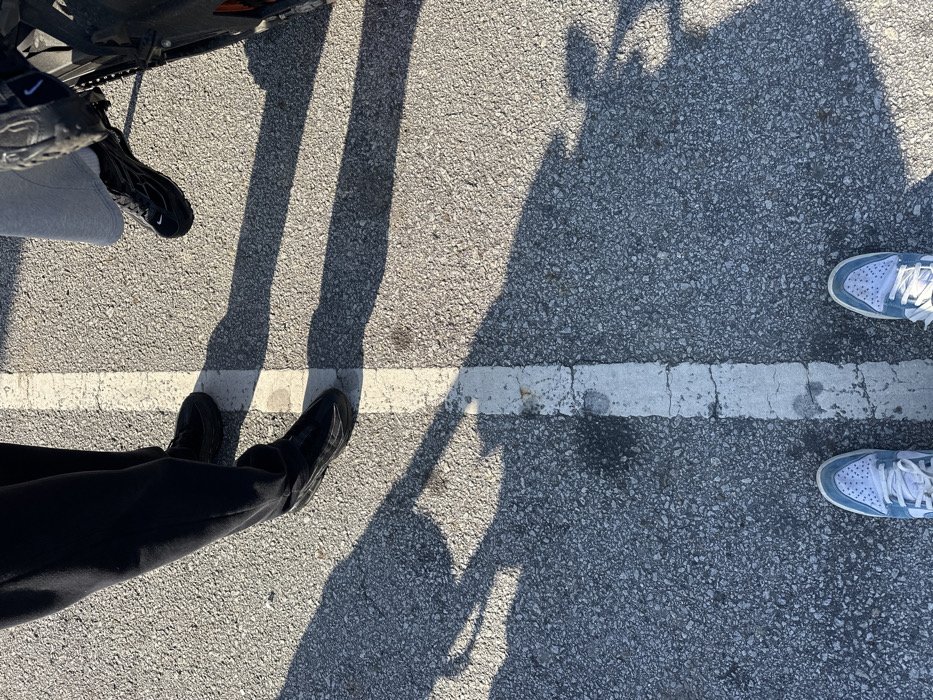 An overhead view depicts three individuals standing on an asphalt surface in Mirandela, Portugal. A worn white line bisects the frame vertically, positioned slightly left of center. To the left of the line, the lower legs of a person wearing dark pants and black shoes are visible. To the right of the line, the lower leg of a second person wearing light grey pants and black sneakers is present, positioned alongside the partial view of a black motorcycle's wheel and frame. At the bottom of the frame, the feet of a third individual, wearing light blue and white sneakers, are positioned on the asphalt, straddling the white line from the perspective of the camera holder. Multiple elongated shadows are cast across the textured grey asphalt surface, indicating a low sun angle.