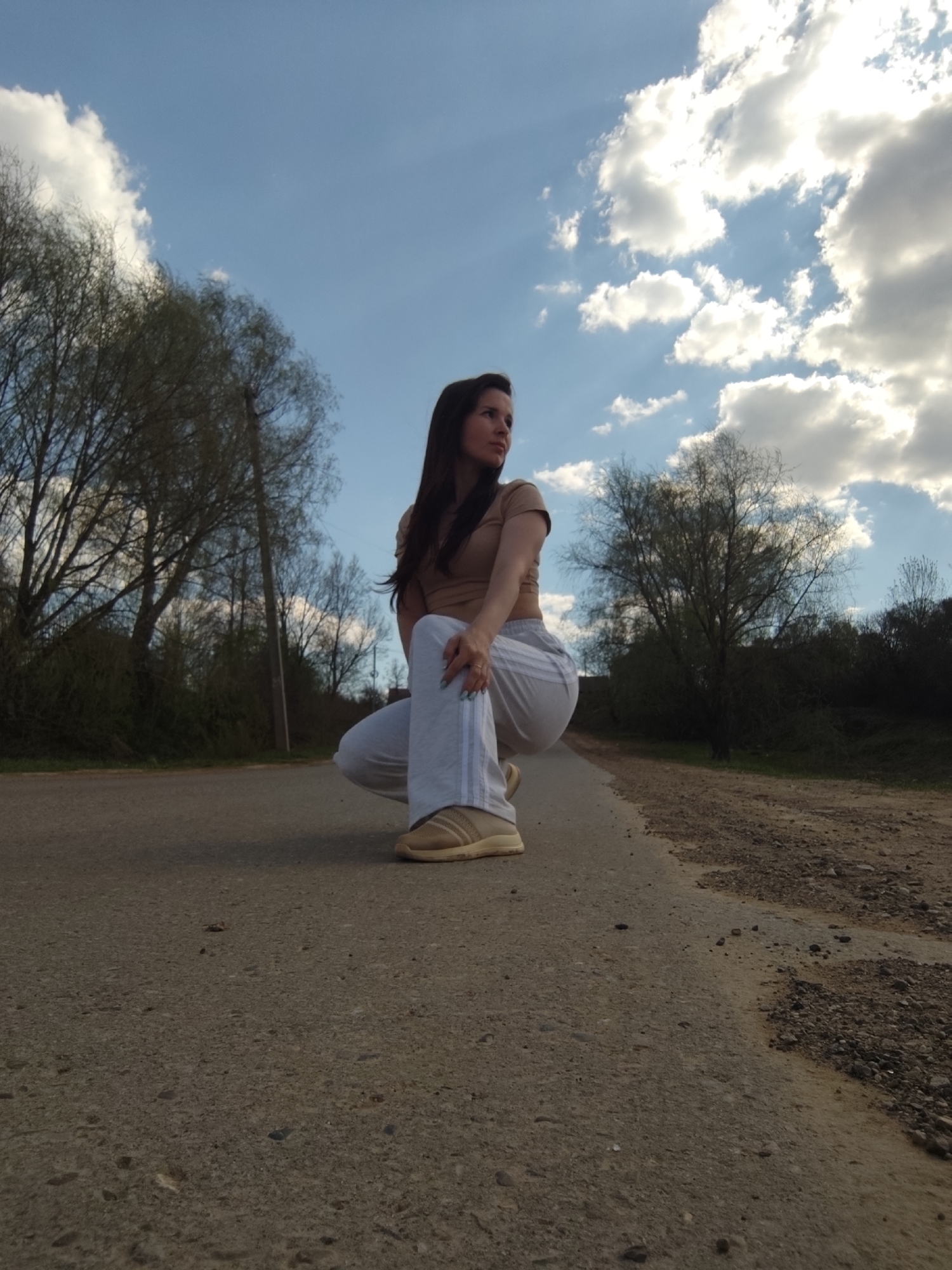 A woman is squatting on a paved road in an outdoor setting in Фоки, Russia. She has long, dark hair and is wearing a light brown t-shirt, light gray sweatpants with stripes, and light brown sneakers. She is looking up and to the right. The road is flanked by trees and bushes. The sky is blue with white clouds.