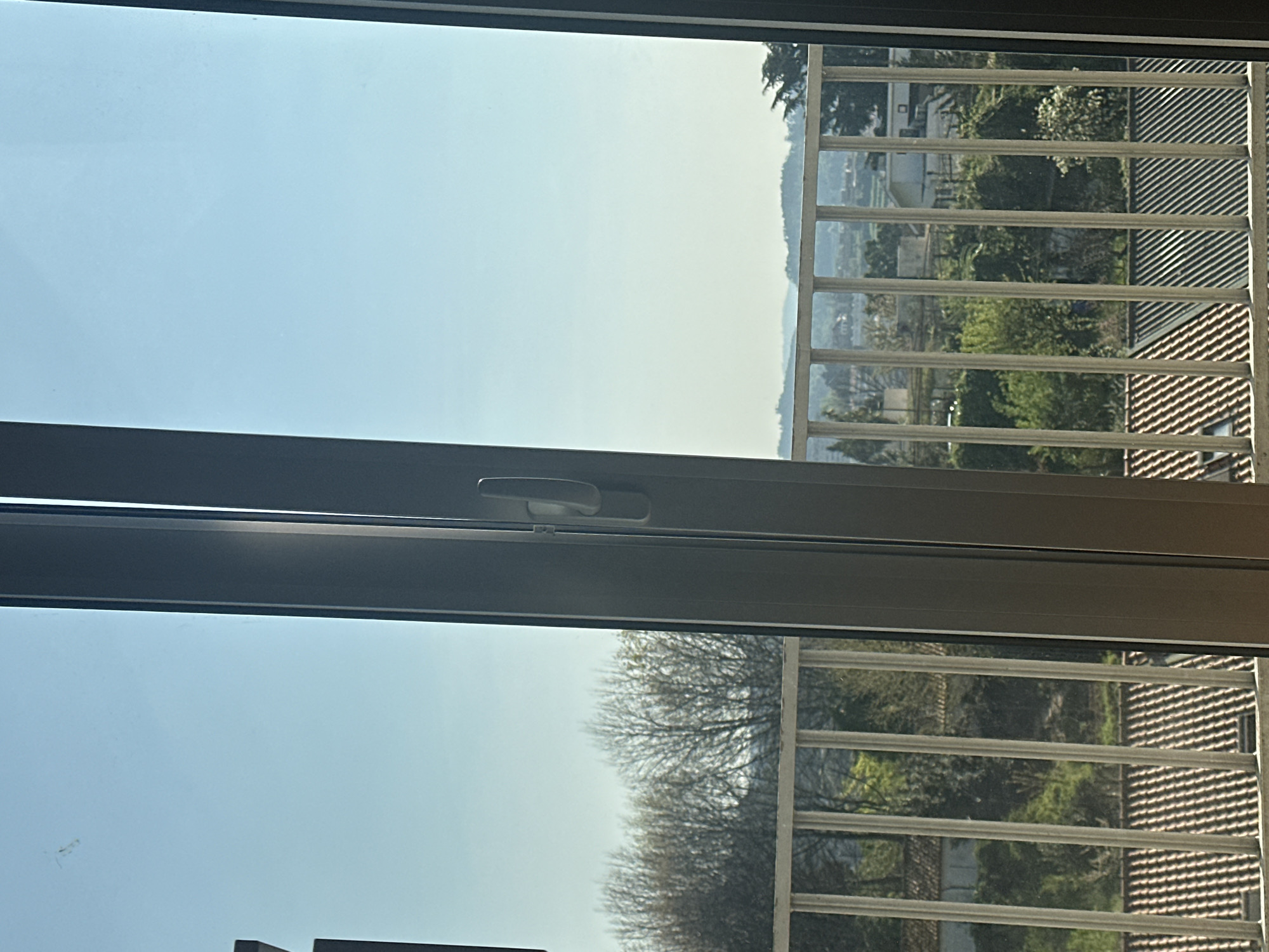 The image depicts an outward view from inside a building, likely through a glass window or balcony door, located in Guimarães, Portugal. A dark-colored vertical window mullion with a light-colored handle occupies the center-right of the frame. Beyond this, a light-colored metal balcony railing with multiple vertical bars extends horizontally across the lower and middle portions of the view.
The scene beyond the railing reveals a varied landscape under a clear, pale blue sky. In the mid-ground, numerous residential rooftops are visible, predominantly featuring reddish-brown terra cotta tiles, with some areas of darker roofing. Green foliage from trees and shrubs is interspersed among these structures and extends into the background. Some deciduous trees with bare branches are also present. The far distance shows a hilly terrain with additional scattered buildings and vegetation, indicating a suburban or semi-rural environment. No people or active interactions are visible. A faint smudge is present on the upper left pane of the glass.
