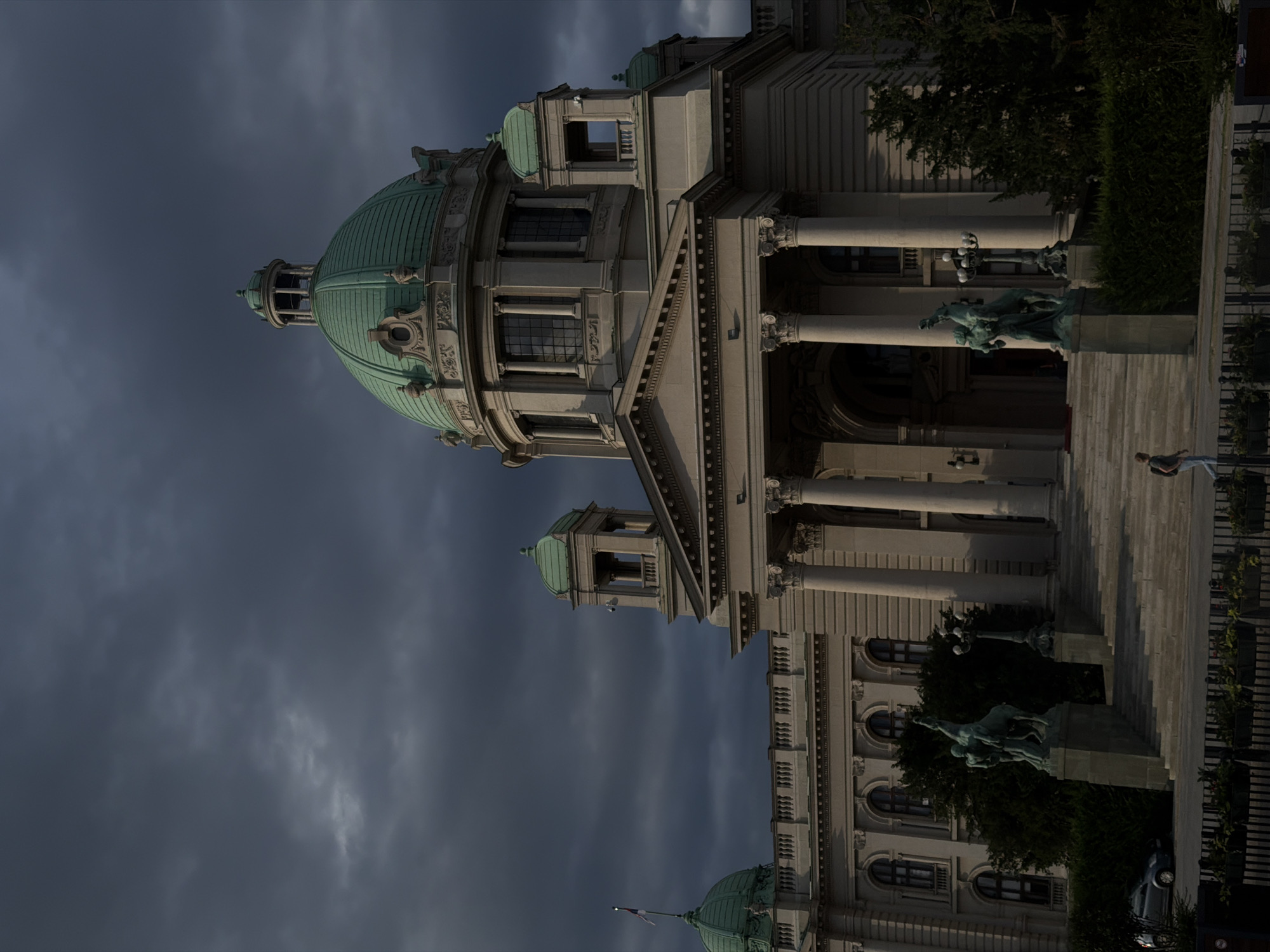 The image shows the National Assembly of Serbia building in Belgrade, Serbia, under a dark, stormy sky. The building features a prominent green dome and classical architecture with columns and statues. In the foreground, a person walks past the building's entrance, while a car is parked to the left. Lush greenery surrounds the building.