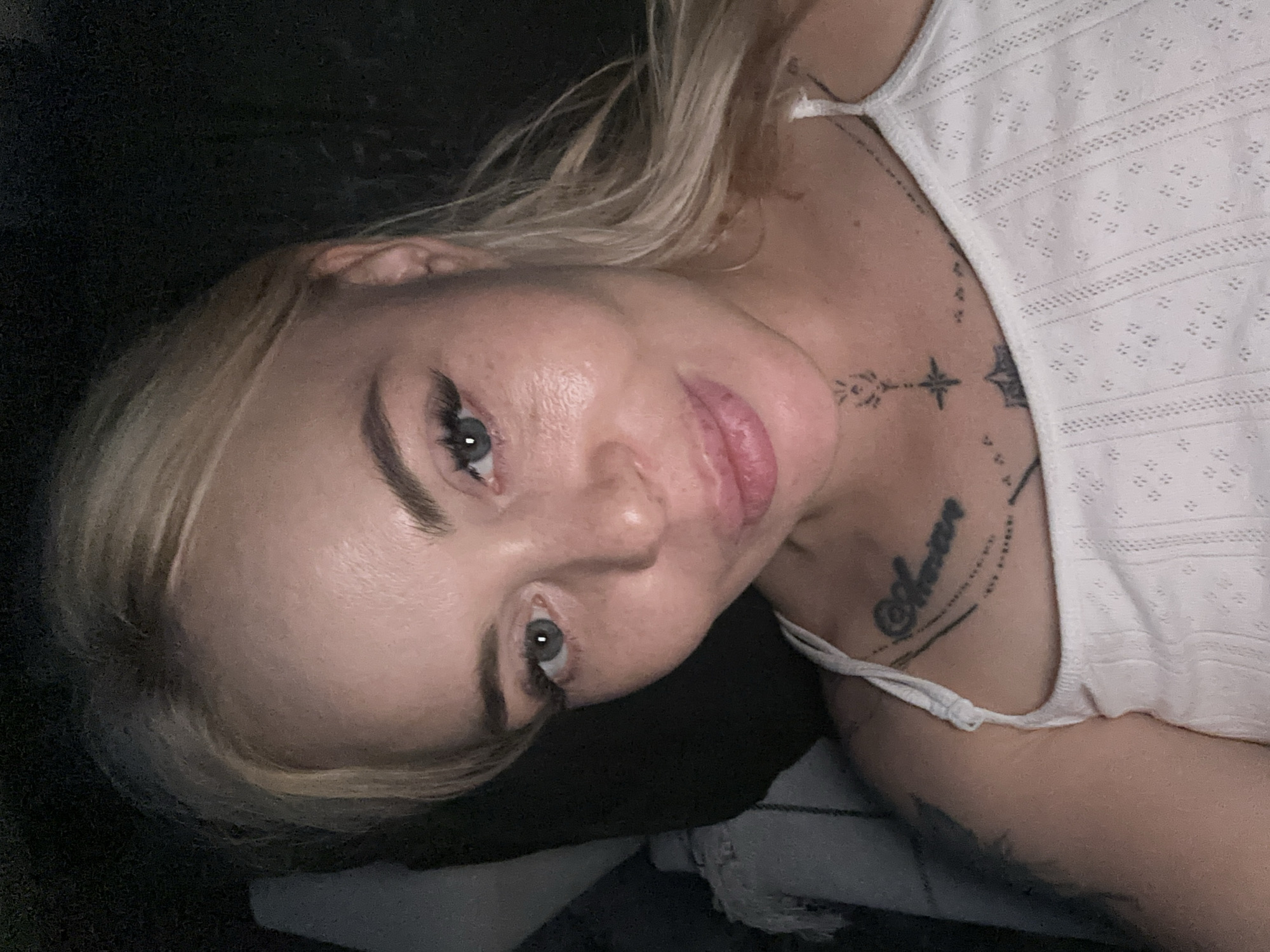 A blonde female subject is depicted in a close-up, head-and-shoulders portrait. She has light skin, blue-grey eyes with prominent eyelashes, and full, slightly parted lips forming a subtle smile. Her blonde hair is parted and frames her face. She wears a white, sleeveless top featuring a textured, eyelet pattern and thin shoulder straps. Tattoos are visible on her upper chest: a script tattoo with decorative lines on the viewer's left side of her chest, a stylized star or compass design centered below her neck with a floral element, and an additional smaller, abstract tattoo to the viewer's right of the central design. The background is dark and out of focus, with a dark, soft object visible behind her head and a lighter, textured fabric partially visible on the viewer's right side of the frame. The lighting suggests a dim indoor setting.