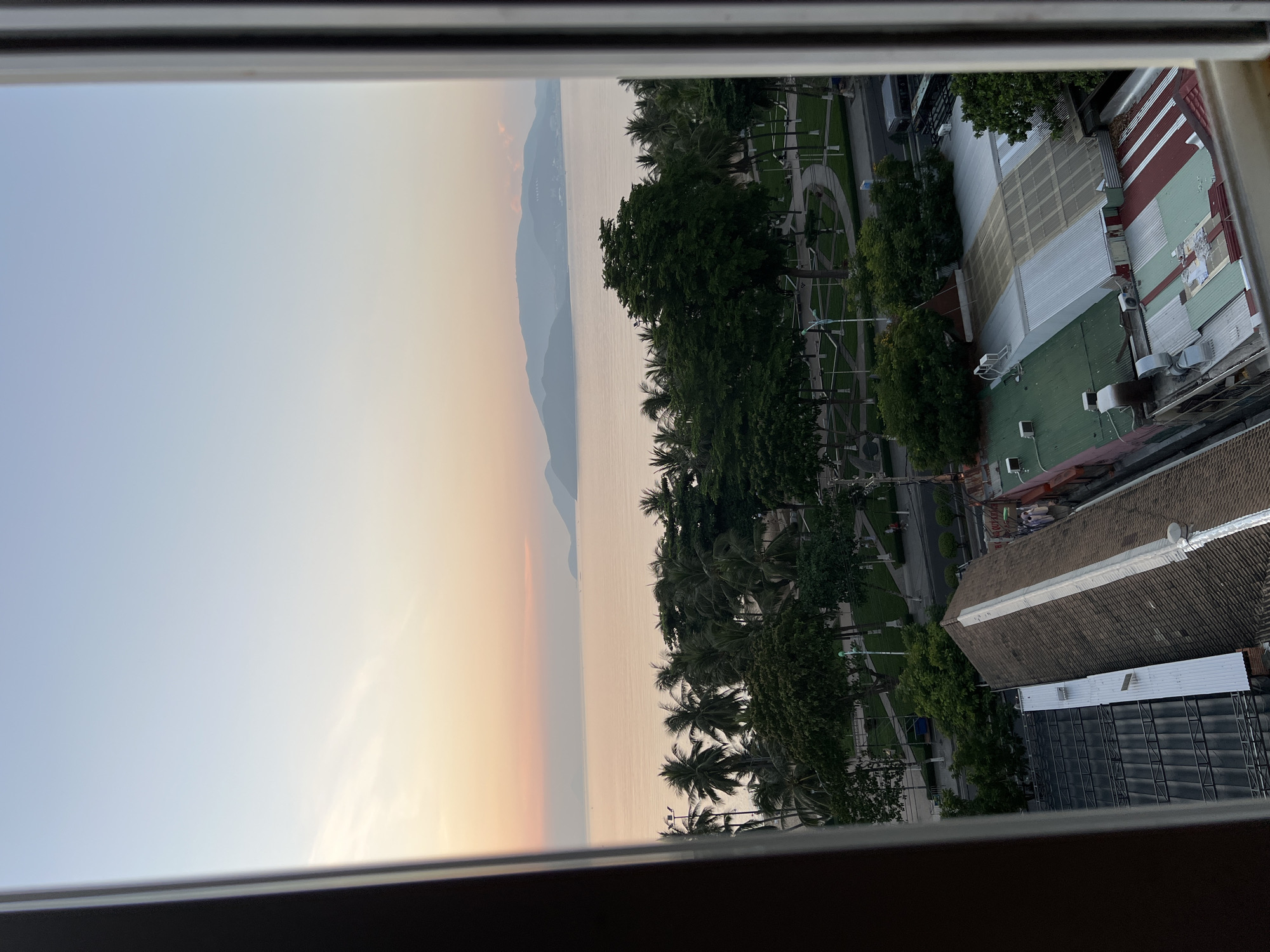 The image captures a view from a window overlooking a coastal city, likely Nha Trang, Vietnam, during sunset. The sky exhibits a soft gradient of pale blue and warm orange hues, with subtle wisps of clouds. The calm sea stretches to the horizon, where hazy mountains are visible. In the foreground, a line of lush palm trees and other dense green foliage separates the beachfront from the urban landscape. Beyond the trees, a park area with winding pathways and streetlights is visible, with a road running alongside it. Further down, a cluster of buildings with diverse rooftops, some made of corrugated metal in various colors and patterns, and others with tiled roofs, dominate the lower portion of the frame. Some buildings have visible air conditioning units. The scene appears peaceful, with no people prominently featured or actively engaged in any discernable actions. The overall impression is that of a tranquil evening in a tropical city.