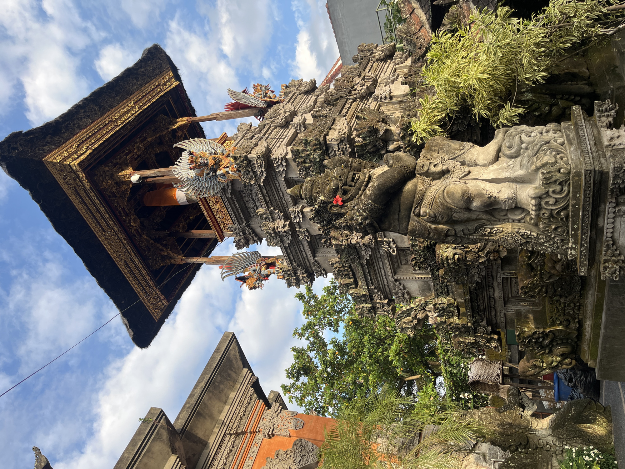 The image captures a temple structure in the Ubud District of Indonesia. The architectural style is distinct, featuring intricate stone carvings and traditional Balinese elements. The structure is adorned with statues, ornate details, and a thatched roof. The sky is visible in the background, with white clouds against a blue backdrop. Vegetation is evident, adding to the natural environment surrounding the temple.