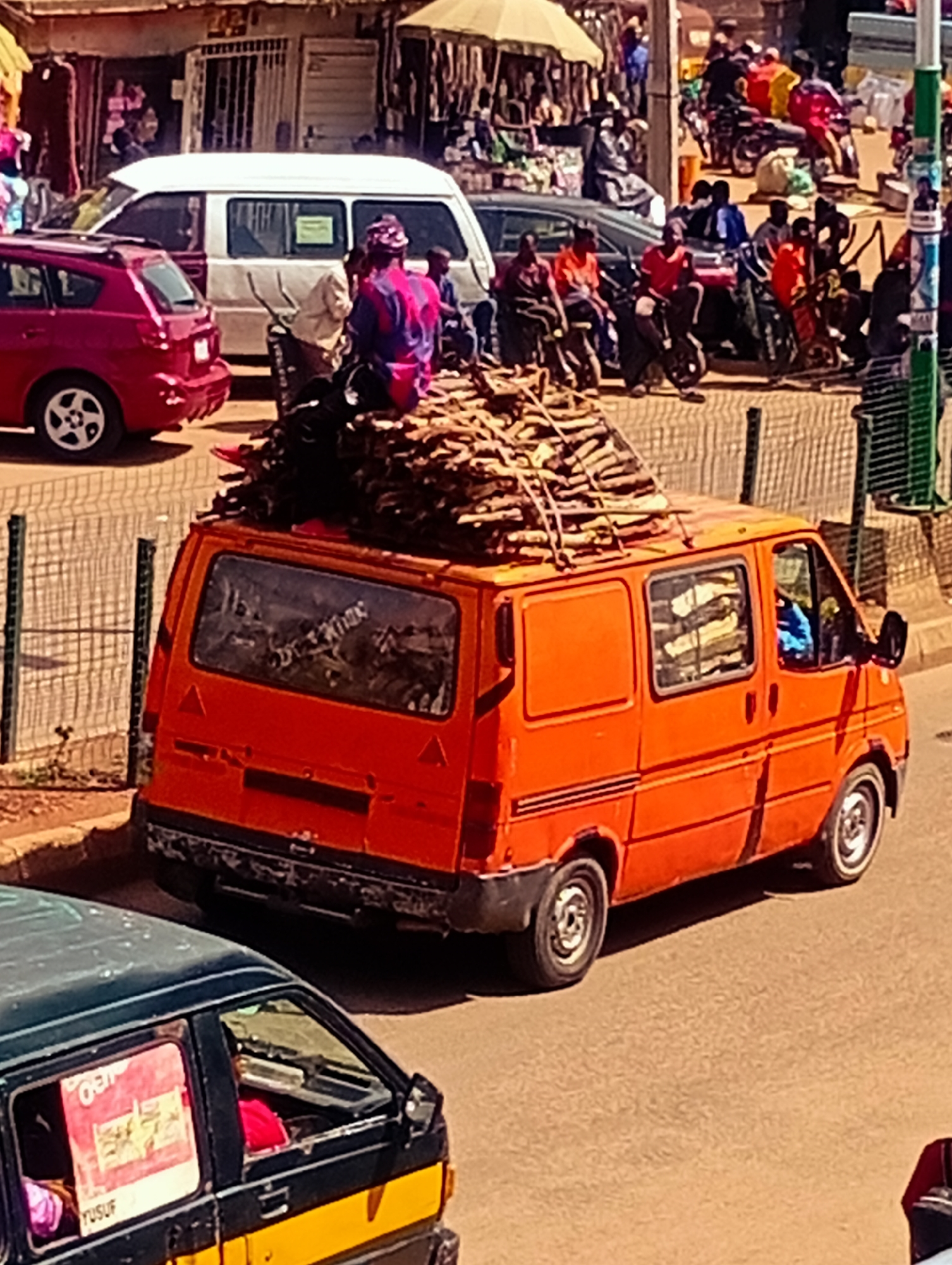 This is an eye-level, medium shot of a bustling street scene in Bukuru, Nigeria, on a sunny day. The foreground is dominated by an orange van, its rear section laden with a substantial pile of firewood. The van's rear window reflects the scene behind it, showing the surrounding market activity. To the left of the van, a red car is parked.
In the middle ground, a white van with tinted windows is also present. People are visible in and around the vehicles, some sitting on the ground, others standing or walking. The background depicts a vibrant market with stalls, awnings, and more people engaged in various activities. Motorcycles and other vehicles are also present, suggesting a busy commercial area. The overall impression is one of lively street commerce.