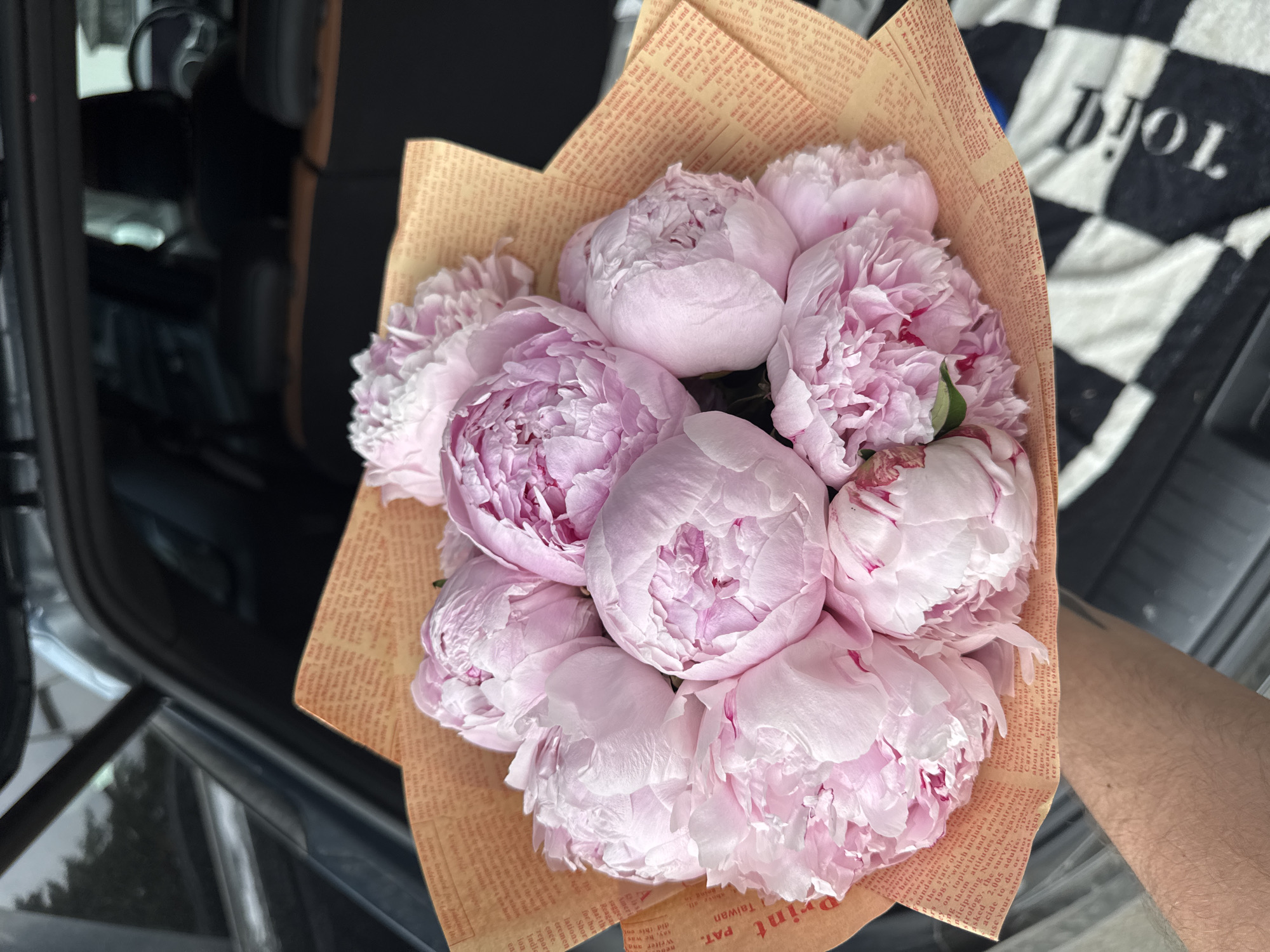 A close-up, overhead shot shows a bouquet of pale pink peonies wrapped in brown paper with text printed on it. The bouquet is held by a person's arm, which is partially visible in the lower left corner. The background suggests the interior of a car, with dark seats and a window. The image is sharply focused on the flowers, with the background out of focus. There is no identifiable location within the city of Almaty, Kazakhstan.