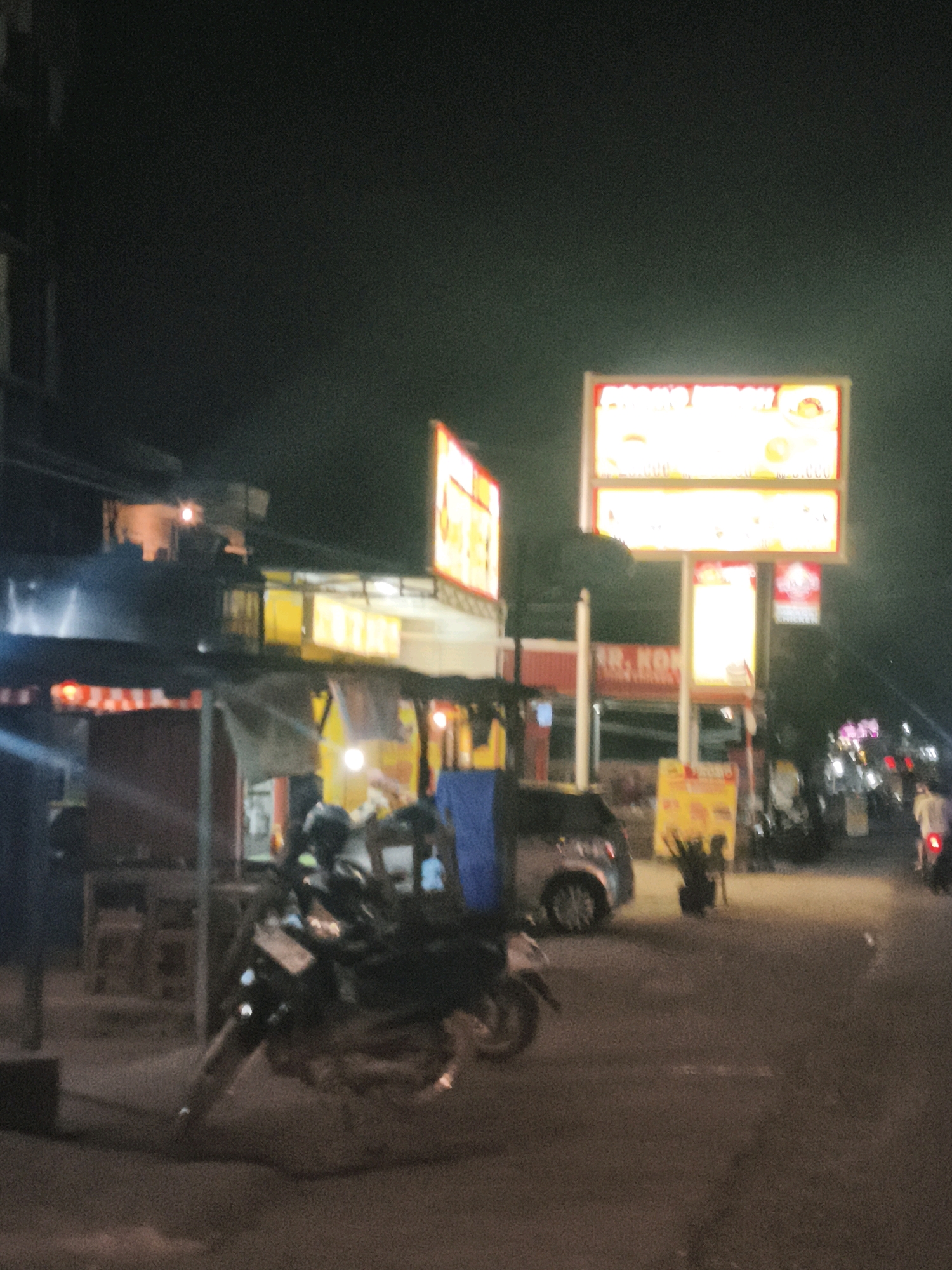 This is a nighttime, slightly out-of-focus photograph of a street scene in Palembang, Indonesia, likely near a commercial area. In the foreground, two motorcycles are parked, one on the left and another slightly further back and to the right. A person wearing a helmet is visible on the motorcycle to the right. Further down the street, to the right, a person on a motorcycle with a lit taillight is moving away from the camera.
The environment includes several structures with illuminated signs. A prominent sign on the right side of the image displays bright yellow text with red accents, appearing to be an advertisement. Below this, another illuminated sign with red and yellow colors is visible, possibly indicating a business name or offering. To the left, a smaller, darker structure with some lighting is present. In the background, there are more buildings and streetlights, contributing to the overall nighttime urban atmosphere. The road surface is asphalt, and there appear to be some street vendors or small shops set up along the side of the road, with tents and tables. The lighting is primarily from artificial sources, creating a contrast between illuminated areas and darker shadows.