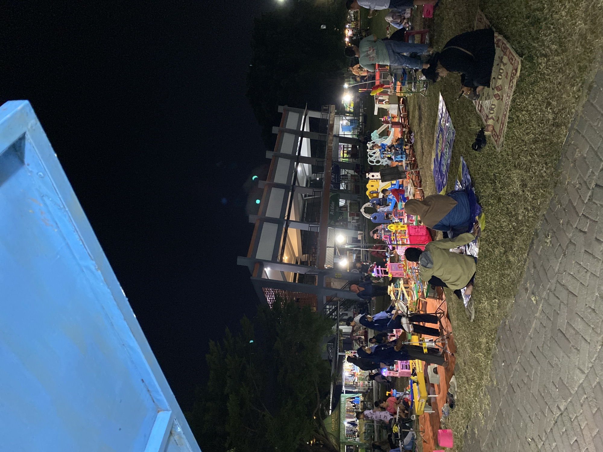 The image depicts an outdoor night market in Madiun, Indonesia, likely held in a public park or open space. The scene is illuminated by artificial lights, creating a warm and lively atmosphere. In the foreground, several people are seated on mats, engaged in various activities such as looking at their phones and interacting with each other. A woman in a black hijab is seated on a patterned rug, engrossed in her mobile phone. Next to her, a person in a green hoodie and blue shorts is sitting on a blanket, facing away from the camera.
The middle ground of the image is filled with various stalls and attractions typical of a night market. There are numerous colorful stalls displaying goods, and a play area with various toys and structures, including a small slide and a toy kitchen. Children and adults are seen browsing the stalls and playing in the designated area. In the background, a modern building with multiple levels stands out, its windows illuminated, suggesting it might be a community center or a commercial establishment. Trees surround the area, adding to the natural ambiance of the location. The overall impression is a vibrant and social gathering, characteristic of night markets in Indonesian cities.