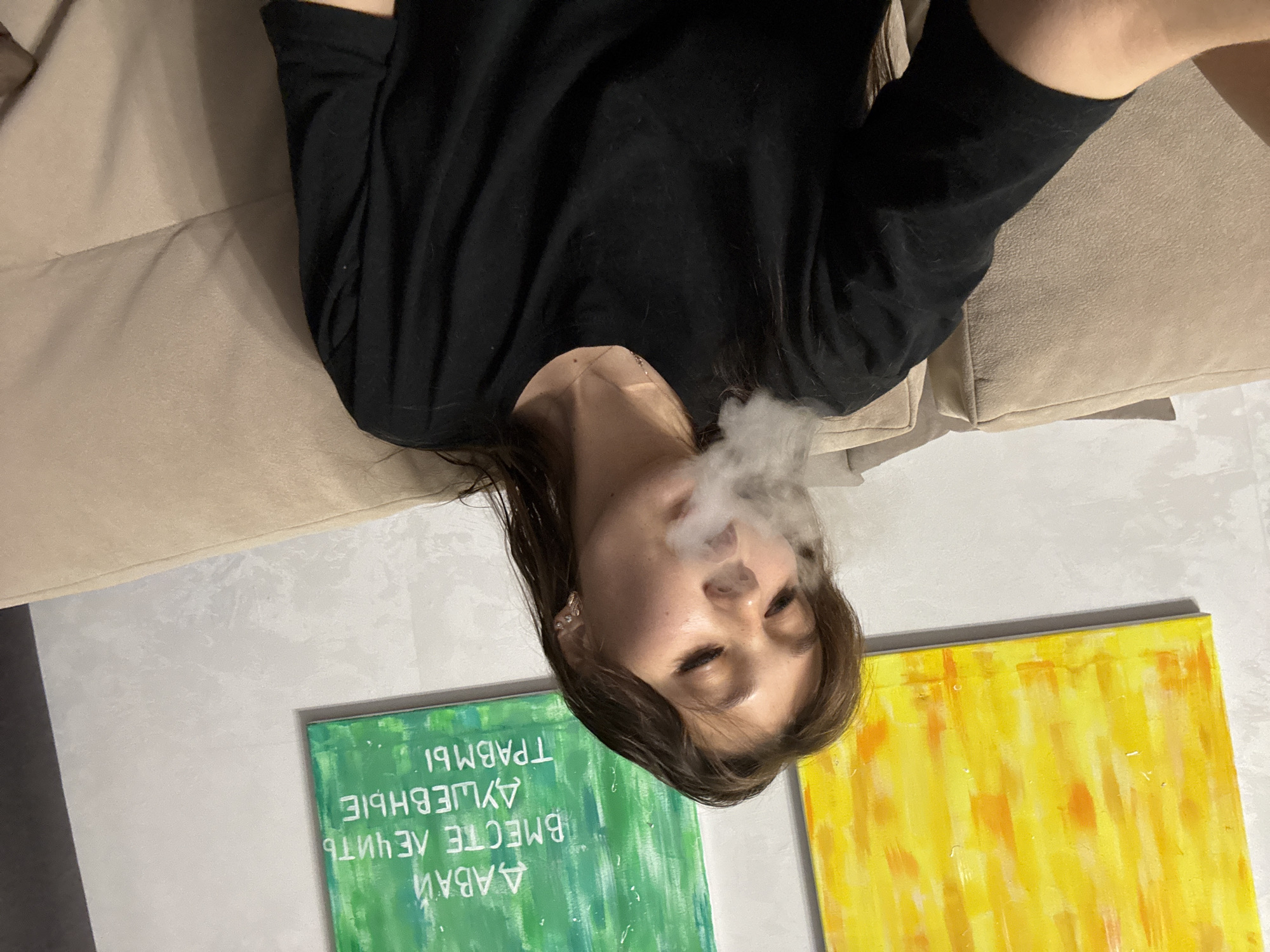 A young woman with brown hair exhales a cloud of vapor while sitting on a beige sofa. She is wearing a black t-shirt and a necklace. Behind her, on the wall, are two paintings: one yellow and orange abstract painting on the left, and a green painting with white Cyrillic text on the right. The text on the green painting reads "ДАВАЙ ВМЕСТЕ ЛЕЧИТЬ ДУШЕВНЫЕ ТРАВМЫ" which translates to "LET'S HEAL EMOTIONAL TRAUMAS TOGETHER". The scene appears to be indoors, possibly a living room, in Novokuznetsk, Russia.