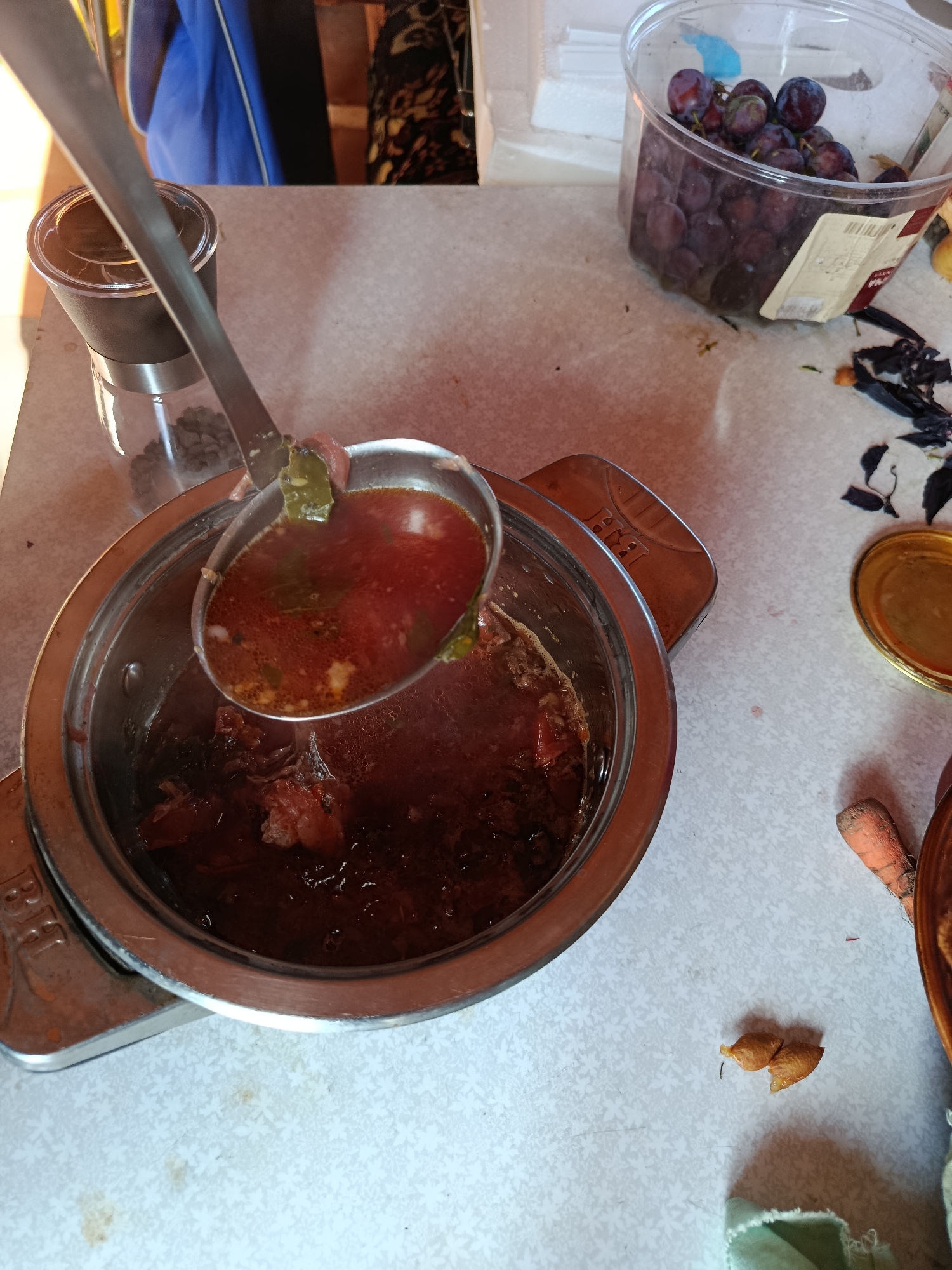 The image shows a close-up overhead view of a pot of stew being served from a metal ladle, on a light-colored table. The stew is a deep red color with visible chunks of meat and vegetables. A clear plastic container filled with dark purple grapes is visible to the right of the pot. There is also a glass grinder with dark contents, possibly coffee beans, to the left of the pot. In the background, a blue and black garment hangs on a rack, and a white object is on a shelf. The table surface has a faint floral pattern and some scattered crumbs. The scene is likely indoors, possibly in a kitchen or dining area.