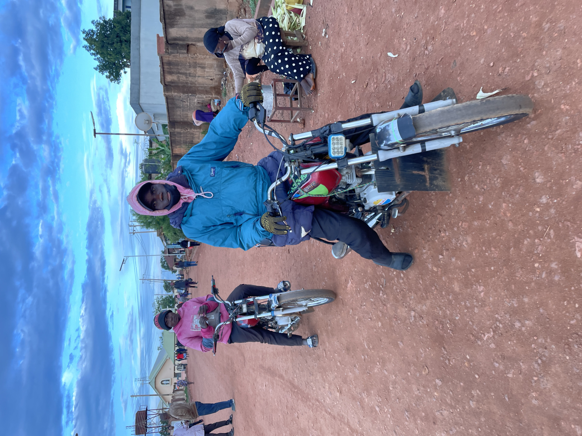 The image captures an outdoor street scene in Gura Top, Nigeria, on a reddish-brown dirt road under an overcast sky with textured grey and white clouds.
In the immediate foreground, a young Black male is seated on a red and chrome Bajaj Boxer motorcycle, facing the viewer. He wears a blue anorak jacket over a pink hoodie, black pants, and yellow-and-black work gloves. A black face mask is pulled down to his chin. The motorcycle features an aftermarket LED light bar on its front and a large, square, black mudguard extension. His left foot, clad in a dark slide sandal, rests on the ground.
Directly behind him, another young Black male is seated on a silver and red motorcycle, also facing forward. He wears a pink hoodie and a dark beanie hat, holding an unidentified object in his hands. He wears patterned slide sandals.
To the mid-right, a Black woman is seated on a low wooden stool, barefoot, wearing a beige top, a patterned apron, a black and white polka-dot skirt, and a dark headscarf. She is holding a mobile phone to her right ear. Adjacent to her, a small wooden stand holds a metallic object, and green leafy produce is visible on the ground.
In the background on the left, a light-colored building with an arched entrance and a statue or emblem above it is visible, positioned next to a metal water tower structure. Electrical poles and overhead wires crisscross the sky. On the right, a low, unpainted concrete wall runs along the road, behind which a building with a flat roof and a satellite dish is partially visible, along with a large green tree. Several other individuals are seen walking or standing further down the street. The road surface is unpaved, uneven, and contains scattered small stones and debris.