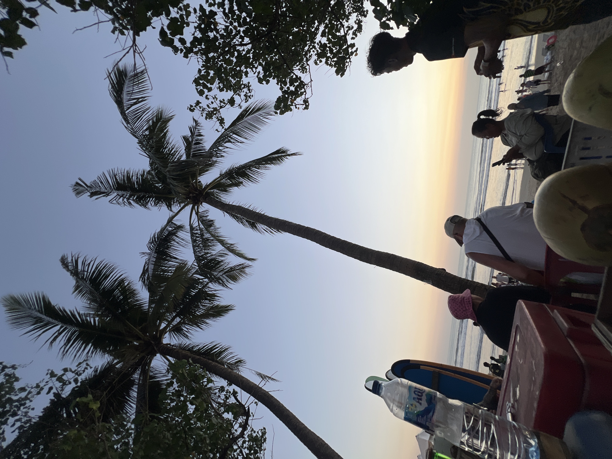 At sunset on Kuta Beach in Bali, Indonesia, a group of people are gathered near the shore. Two palm trees dominate the foreground, their fronds silhouetted against the pale orange and blue sky. In the immediate foreground, a red cooler box sits with a bottle of AquA water and two surfboards resting against it.
To the right, a young man in a black t-shirt and patterned shorts stands talking to another person. To the left, a woman wearing a pink hat and black shirt is seated, facing away from the camera, while a person in a white t-shirt and cap sits beside her. Further back, on the beach, numerous people are visible, some walking along the shoreline and others interacting with the water. The ocean waves are gentle, and the sand appears wet from the receding tide. The overall atmosphere suggests a relaxed evening on a popular tourist beach.