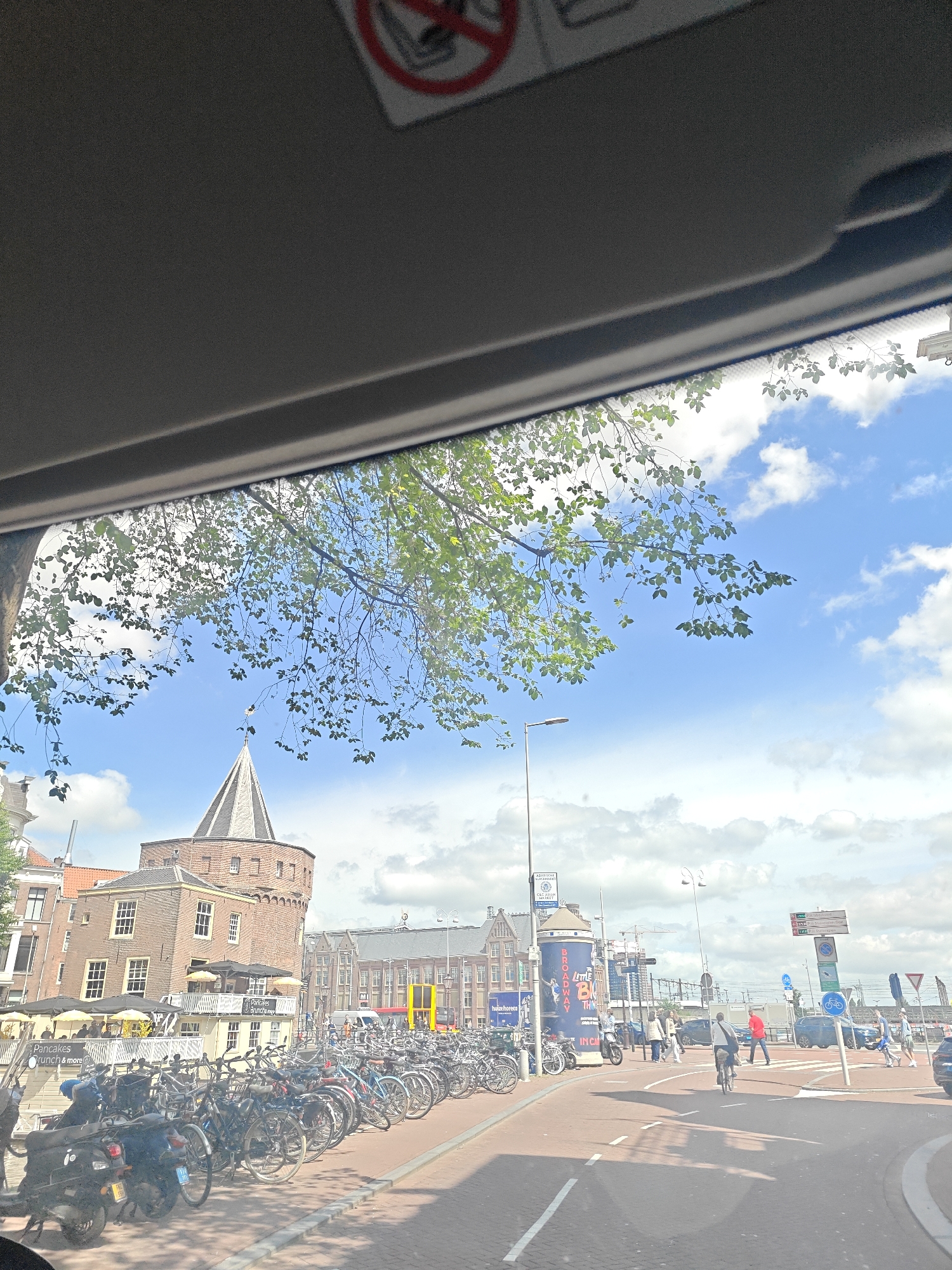 The image captures a street scene in Amsterdam, Netherlands, likely viewed from inside a vehicle. In the foreground, a sun visor is visible at the top, partially obscuring the sky. Below the visor, lush green tree branches hang into the frame, with the blue sky and scattered white clouds forming the background.

Dominating the middle ground is a distinctive brick tower with a pointed, dark-colored roof, resembling a historical landmark or gatehouse. Adjacent to it, a multi-story brick building with numerous windows and awnings suggests a commercial or residential area. A row of parked bicycles, a common sight in Amsterdam, stretches along the sidewalk in front of this building, with a few motorcycles also present. Several small yellow umbrellas are open on a patio area, indicating an outdoor seating space for a cafe or restaurant. One sign visible on the building reads "Pancakes Brunch & more."

Further into the background, more buildings of varying architectural styles are visible, some with prominent yellow accents. A large blue cylindrical billboard is also present, featuring text including "BROADWAY" and "LITTLE SHOP OF HORRORS." Traffic signals and street signs are dotted throughout the scene.

In the distance, traffic can be seen on a road, including cars and people cycling and walking. A pedestrian crossing is marked on the road where individuals are navigating the intersection. The overall impression is of a bustling urban environment on a bright, partly cloudy day, characteristic of Amsterdam's city life.