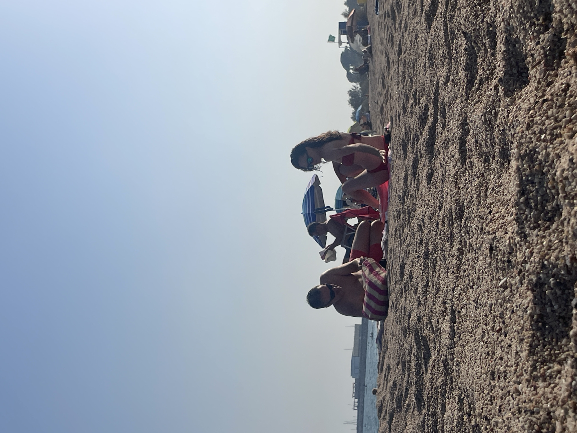 At a beach in Arenys de Mar, Spain, three people are relaxing on the sand. A man in the foreground is lying down, wearing sunglasses and a striped bag. Beside him, another person is also lying down, partially obscured by an umbrella. To the right, a woman in a red bikini is sitting up and looking to the side. Several beach umbrellas and other beachgoers are visible in the background, along with the sea and a distant shoreline with buildings. The sky is clear and blue.