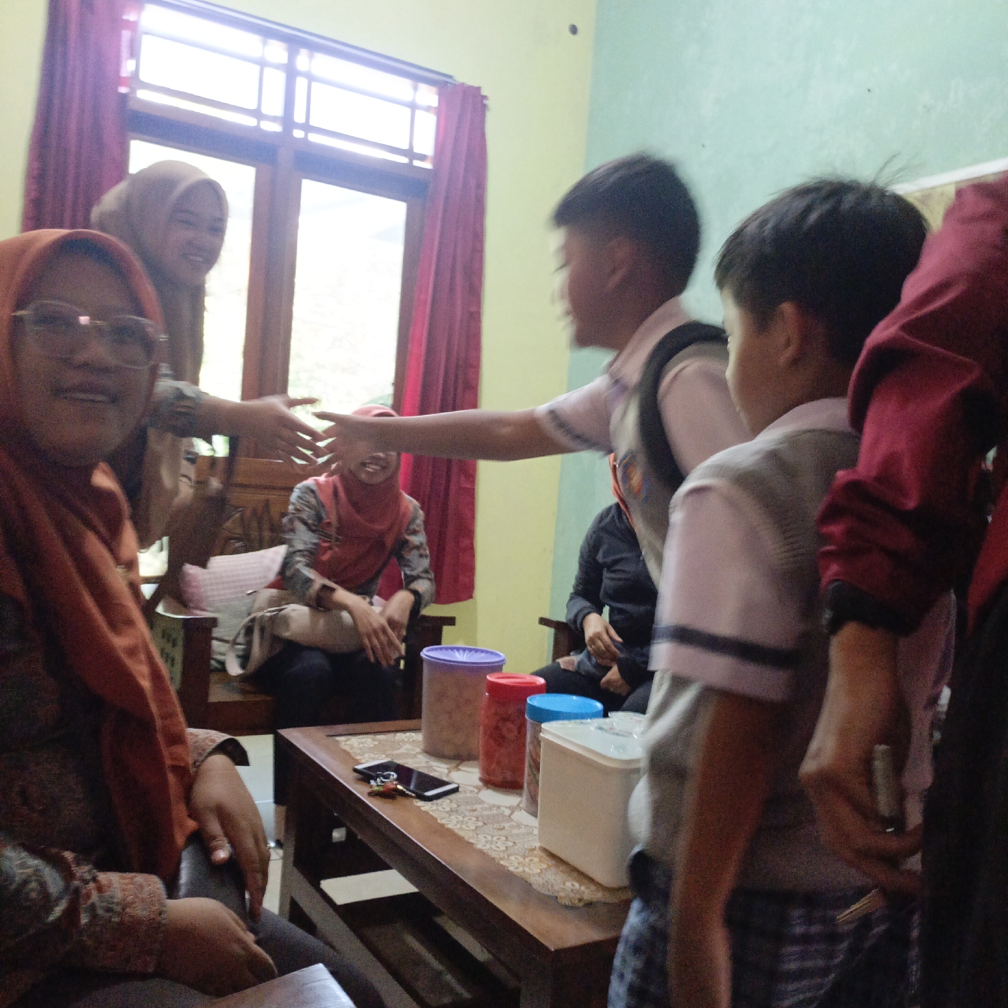 In Salatiga, Indonesia, a group of individuals is gathered indoors, likely in a home or community space. The scene suggests a social interaction or gathering.
In the foreground, a woman wearing an orange hijab and glasses is visible, looking towards the camera with a smile. To her left, another woman in a tan hijab is also smiling and looking to the side. Their hands are near each other, potentially in a gesture of greeting or conversation.
In the middle ground, two boys, possibly children or adolescents, are present. One boy, wearing a white shirt with dark stripes, is in motion, extending his arm. Another boy, in a grey vest, is also facing away from the camera.
In the background, another woman in a reddish-orange hijab is seated, with her hands resting in her lap. The environment appears to be a room with a window covered by burgundy curtains, and a wall painted in a light green hue. A wooden table is in the center of the frame, holding several containers, a smartphone, and keys. The lighting suggests it is daytime, with light coming in from the window. The overall atmosphere seems casual and friendly.