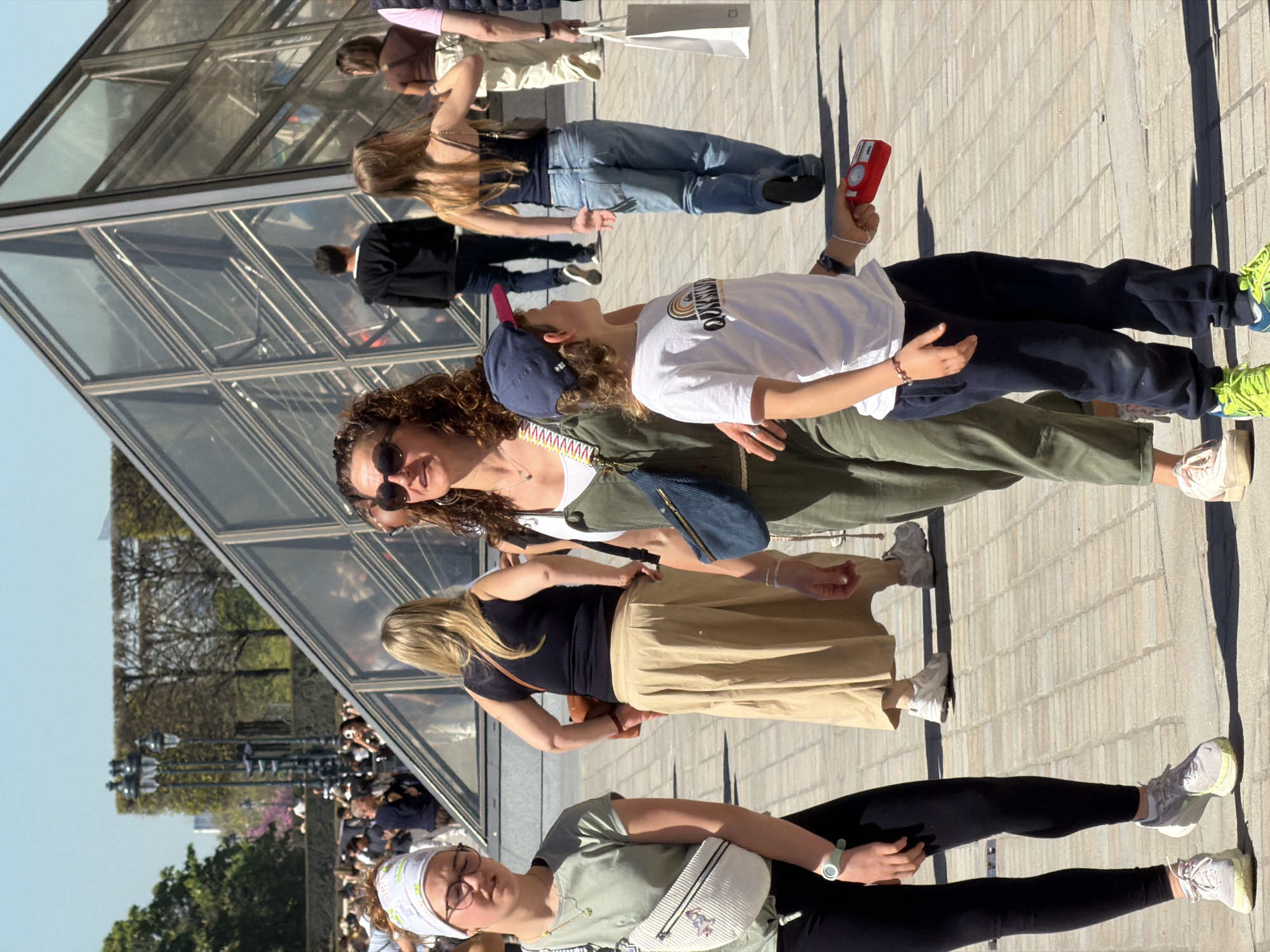 The image captures a group of people standing in front of the Louvre Pyramid in Paris, France. There are at least six individuals visible, including a person wearing sunglasses, an olive green jumpsuit, and a denim bag; a person wearing a white shirt and blue baseball cap, holding a red object; a person wearing a black top and a tan skirt; and a person wearing a light green shirt, black leggings, and a white crossbody bag with a patterned headscarf. The Louvre Pyramid’s glass and metal structure is visible in the background, reflecting the surroundings. There is a crowd of people and trees in the far background. The ground is paved with rectangular stones.