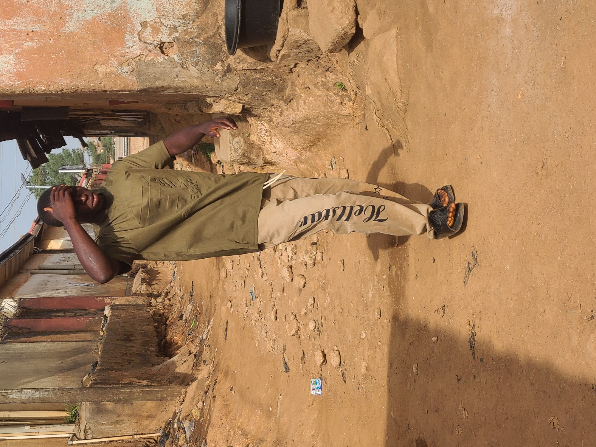 A medium-shot captures a man outdoors. He is wearing an olive-green shirt and khaki pants, with the word "Hollstar" printed on the side of his pants. He also wears dark sandals. His right hand is on his forehead. The environment appears to be in the city of Jos, Nigeria. The background consists of a weathered building and a dirt road or path. There is a black bucket on top of a rock in the background.