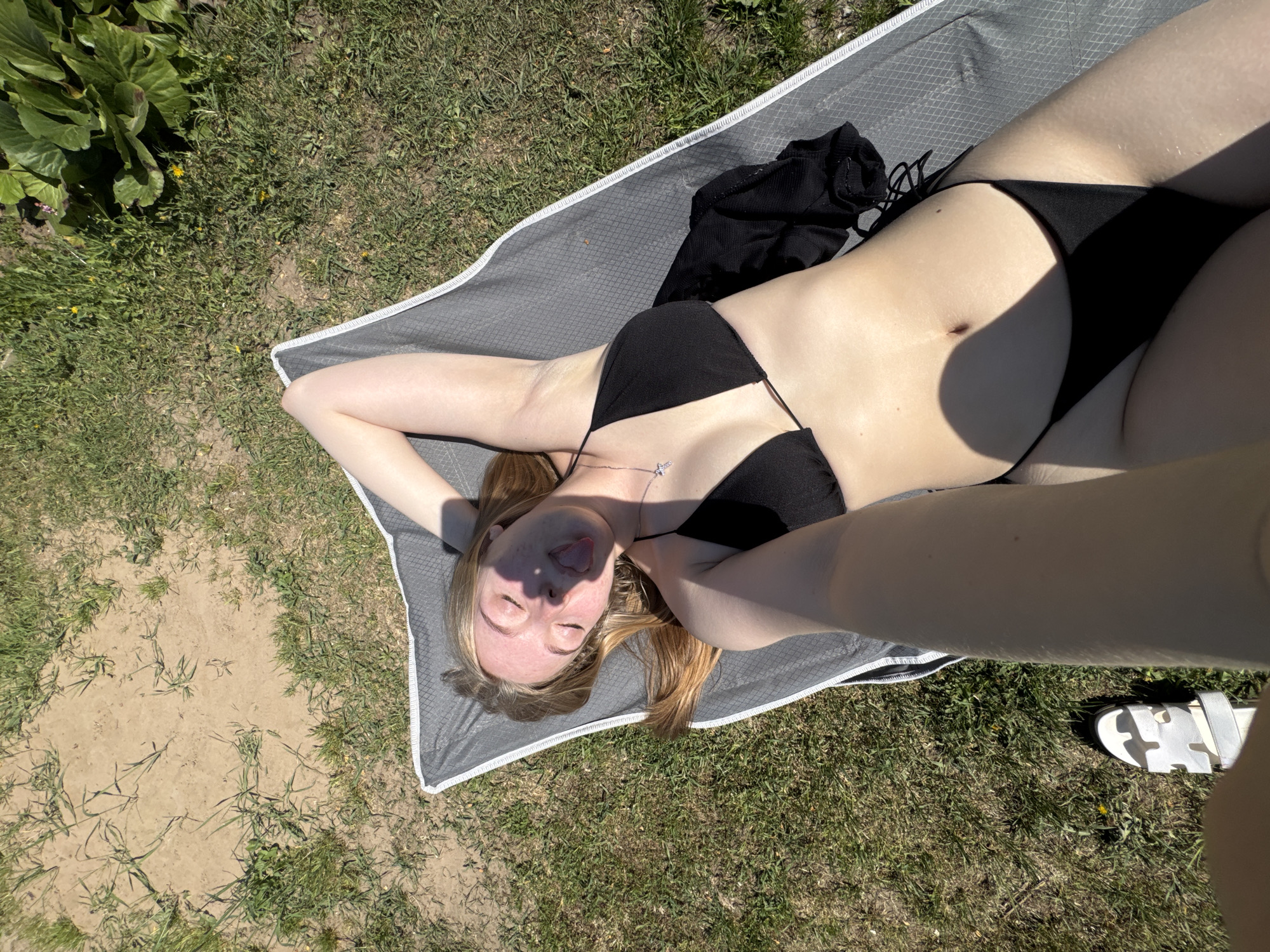 A fair-skinned young woman lies on a gray blanket on grass. She is wearing a black bikini. Her eyes are closed, and she is sticking her tongue out. A black garment is lying near her on the blanket. White sandals are visible at the lower right corner of the image. The location is outdoors, with grass and some plants visible. The city of Zhigalki, Russia, is not discernable in the image.