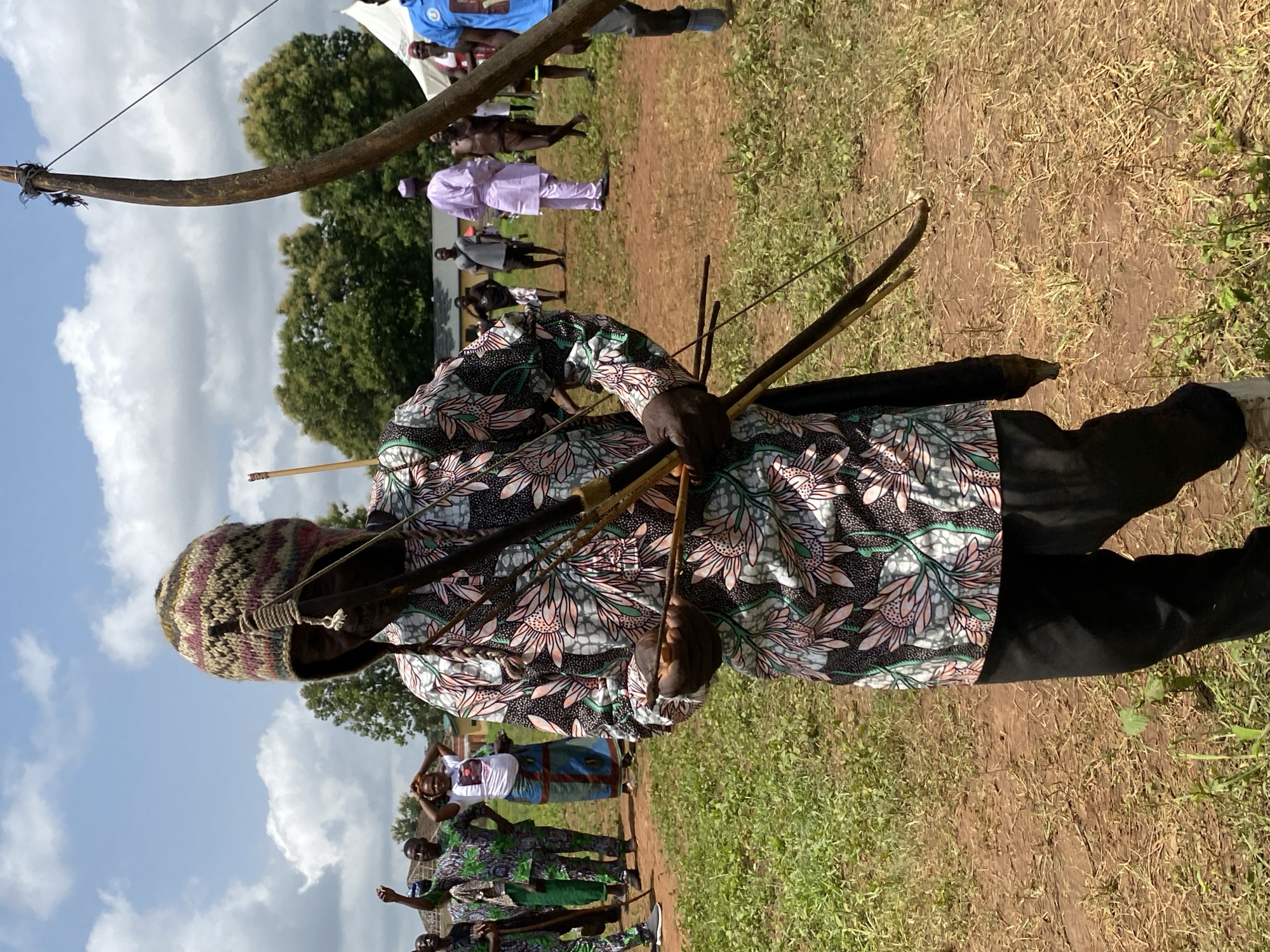 An African man in Maigizo, Nigeria, stands on a patch of dry dirt and grass holding a bow and arrows. He wears a patterned floral shirt, black pants, and a knit hat. Behind him are other people and a wooden structure. The sky is visible with some clouds.