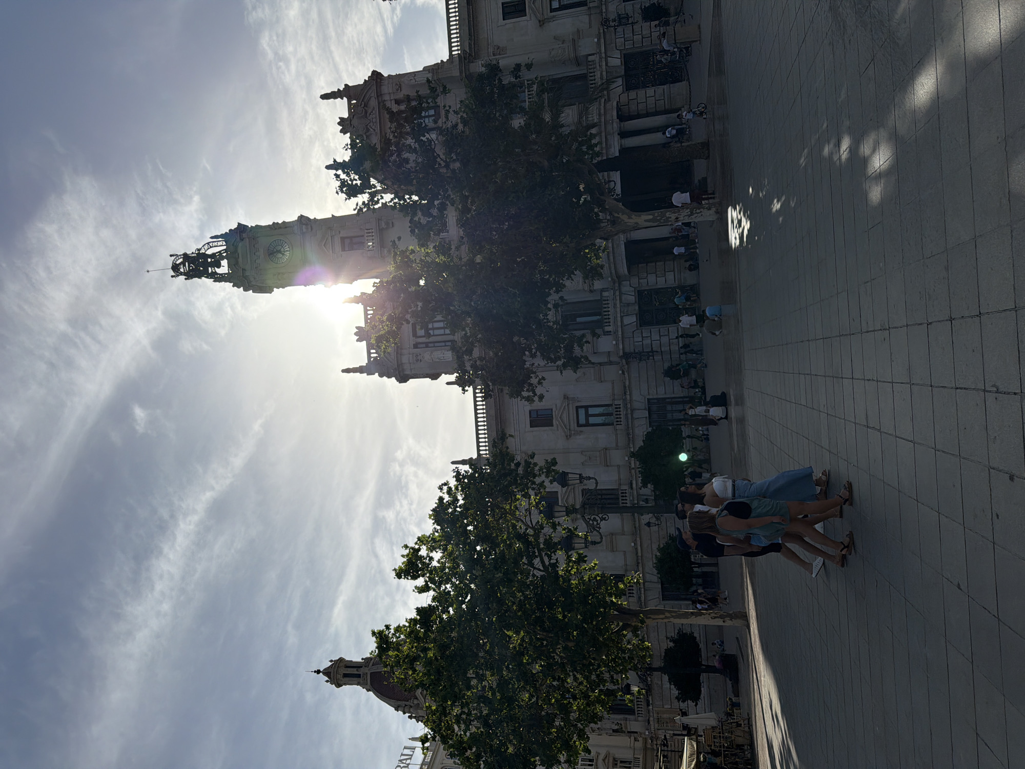 In a public square, a group of individuals walk across a tiled pavement. A large, ornate building with a prominent clock tower stands in the background, partially obscured by trees. The sun is bright, casting long shadows and creating lens flare effects in the sky. The architecture suggests a European city.