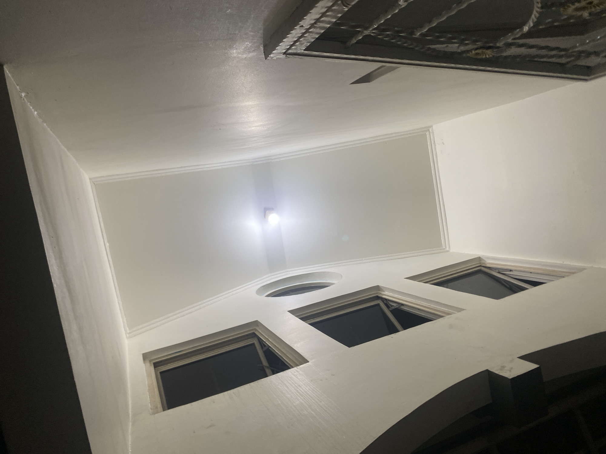 An indoor, low-angle, wide shot shows a courtyard from below, possibly in Dukuh, Indonesia. The walls and ceiling are painted white and show a slight sheen. The ceiling has a recessed rectangular section with decorative molding. A light fixture is centered in the recessed ceiling, emitting a bright light. On the left wall, there are several dark-windowed openings, including one circular opening. On the right, a dark, ornate metal grate is visible. The overall impression is of a clean, well-lit, modern interior space.