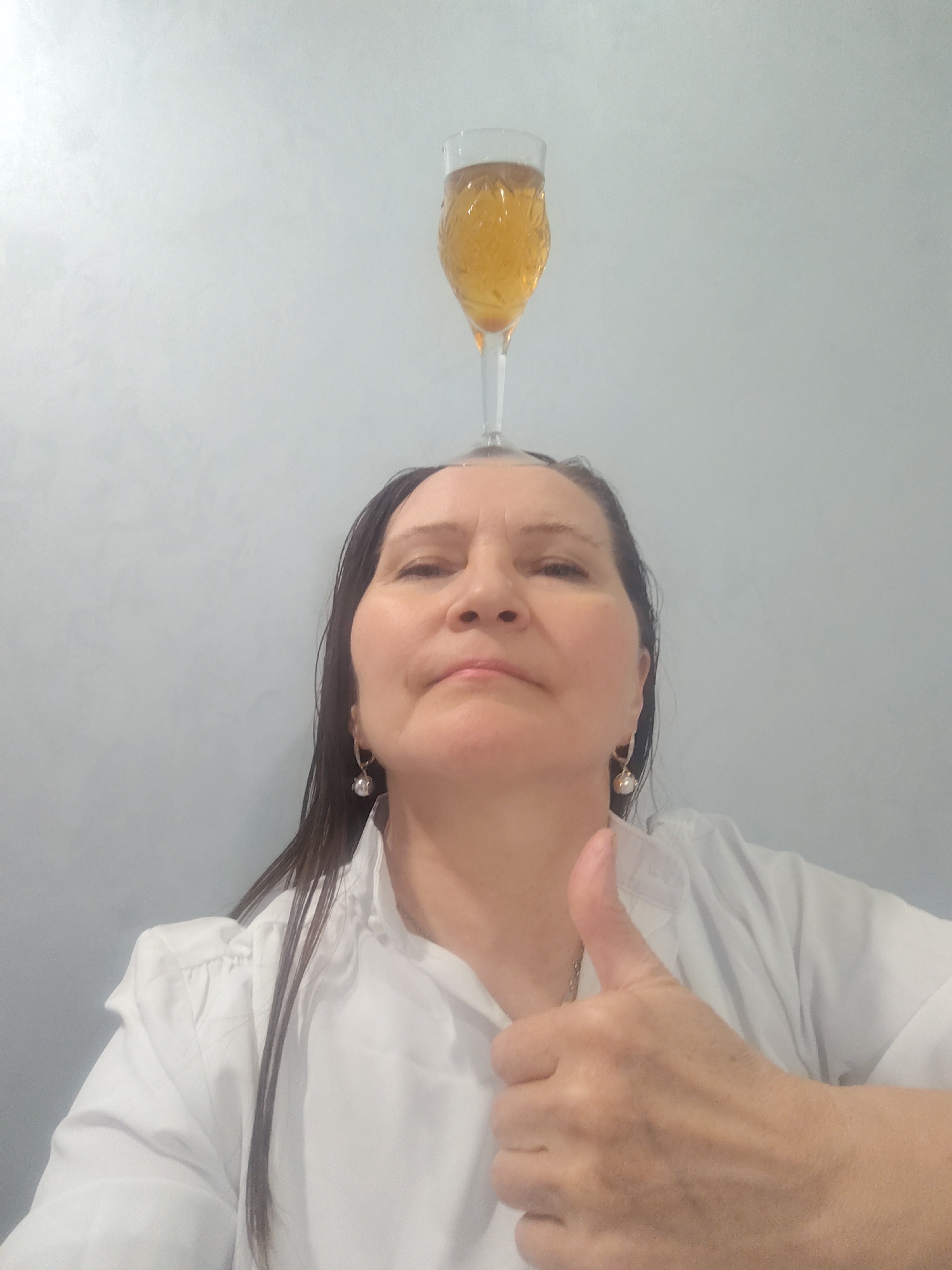A medium shot shows a woman posing with a glass of beer or cider balanced on her head. She gives a thumbs-up with her right hand. The woman has medium-length, dark hair that appears wet. She wears a white button-down shirt and small, round earrings. The background is a plain, light-colored wall. There is no information suggesting the location is Sarapul, Russia.