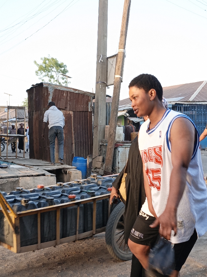 The image depicts an outdoor street scene in Gwagwarwa, Nigeria. In the immediate foreground on the right, a person with short dark hair, wearing a white sleeveless basketball jersey with red "INGS 55" text and an NBA logo, and black shorts, walks from right to left. A black garment is draped over their left arm, and a dark object is held in their left hand.
Immediately to the left of this person, a wooden pushcart is positioned, laden with approximately eighteen dark-colored rectangular containers, each featuring an orange cap. In the mid-ground, a male subject wearing a light grey hoodie and dark trousers is observed from behind, stepping onto a raised wooden platform that leads to a structure made of weathered, rusted corrugated metal sheets. A blue plastic barrel is situated near the base of this structure.
Two utility poles, one concrete and one wooden, stand vertically in the center-right, supporting overhead wires. The background features various buildings with corrugated metal roofs, additional pedestrians, and a parked motorcycle on the far left. The ground throughout the scene is unpaved dirt. The sky is clear and bright.