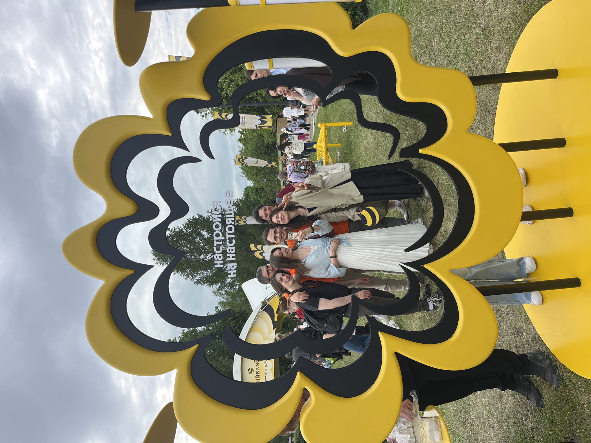 In a park setting, likely in Moscow, Russia, a group of six individuals are posing for a photograph within the frame of a large, artistic installation shaped like a stylized flower. The installation is primarily yellow with black accents and appears to have multiple layers or segments. The people are standing together, with some holding up a phone to take a picture. In the background, other people and structures are visible, suggesting a public event or gathering. The sky is overcast with clouds.