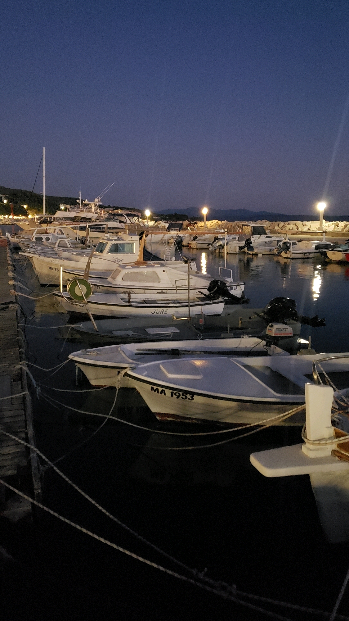 A night-time, medium-shot, vertical photograph captures a harbor in Općina Tučepi, Croatia, with several white boats docked. The sky is a deep twilight blue, with a few distant lights visible along the shoreline and on the water. The boats are lined up next to a wooden pier, with white ropes securing them to the dock. Some of the boats have outboard motors attached to their sterns. One boat in the foreground has "MA 1953" painted on its hull. The water is dark and reflects the lights from the harbor. The overall scene is peaceful and serene.