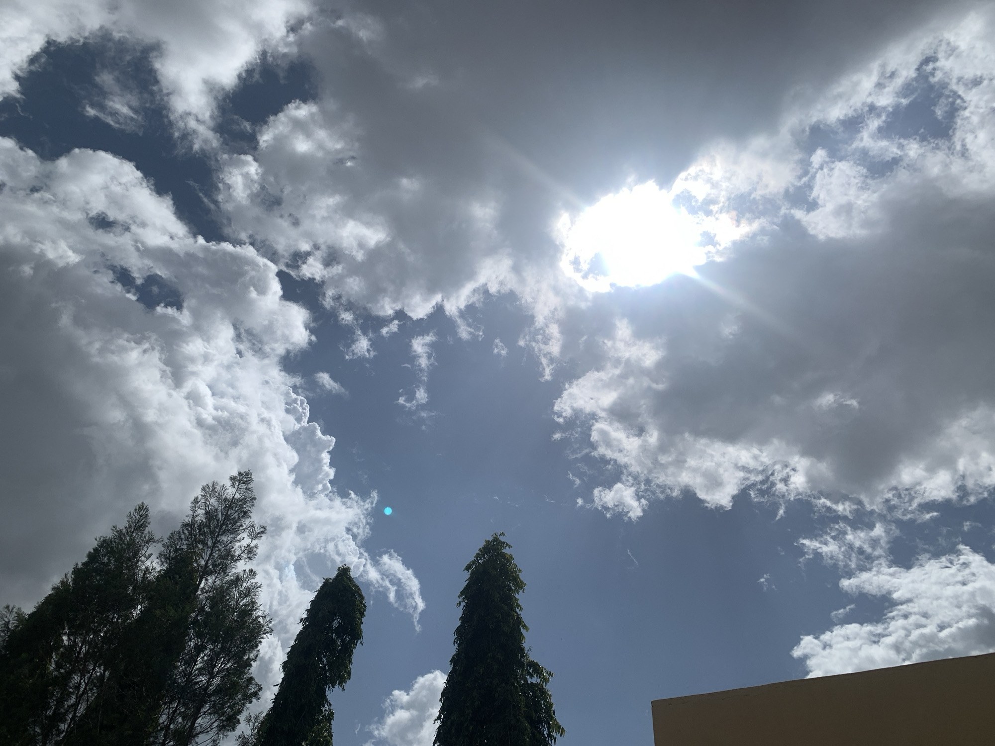 The image depicts a partly cloudy sky over Kuru Karama, Nigeria. The sun is partially obscured by dense clouds, casting rays of light through the gaps. Tall, dark green trees are visible in the foreground, silhouetted against the brighter sky. A portion of a light-colored building is visible in the bottom right corner. The overall scene suggests a dynamic weather pattern, with areas of bright sunlight and shadow.
