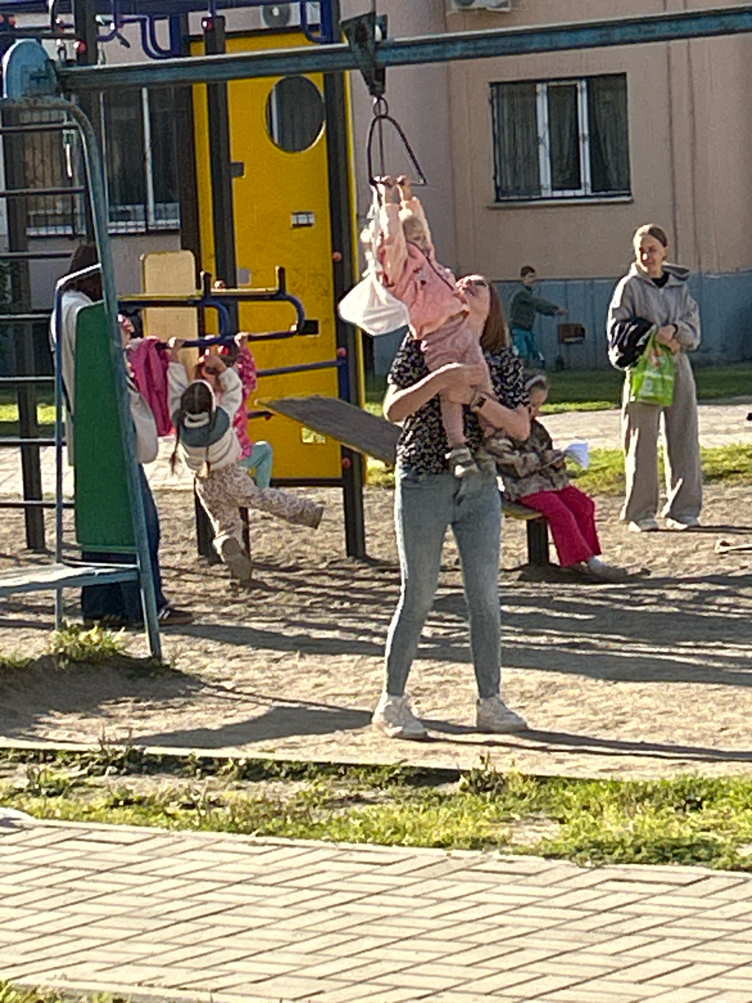 A group of people, including children and adults, are gathered at a playground in Tomsk, Russia. A woman is holding a toddler in her arms, while another child swings on a bar above. In the background, other individuals are present, and a building with windows and a playground structure are visible. The ground is covered with sand and paving stones.