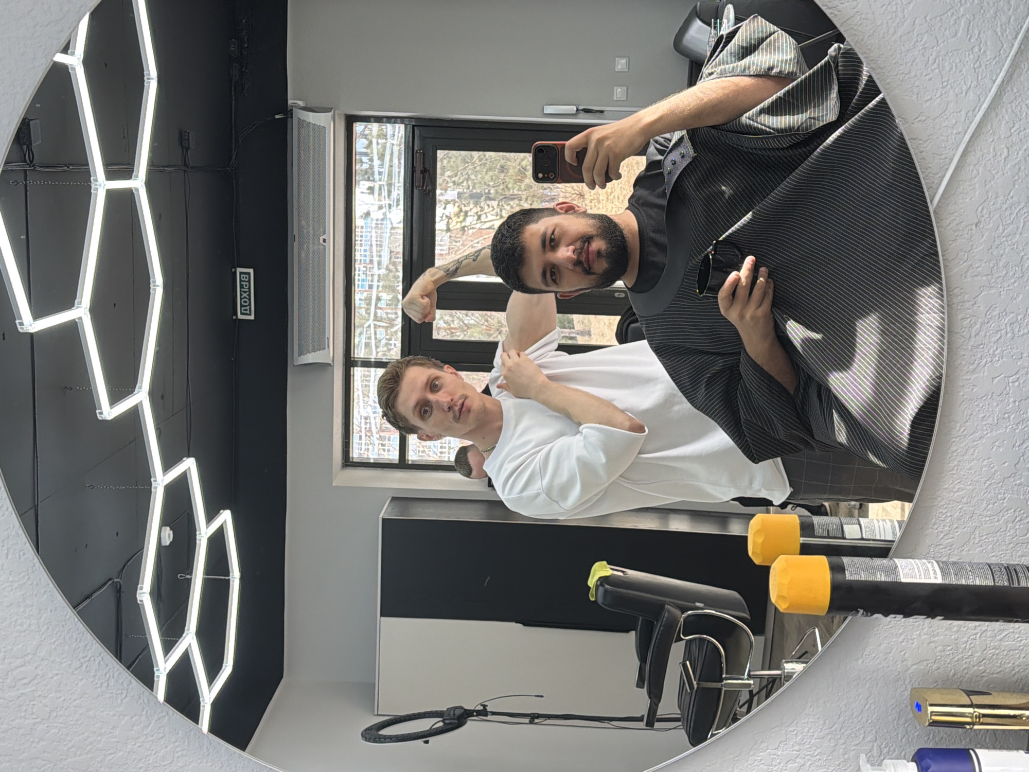 A man takes a selfie in a circular mirror in a barber shop. The man in the reflection on the right holds a phone and looks at the camera. He has short dark hair, a beard, and is wearing a dark-striped barber cape. The man on the left flexes his bicep. He has short brown hair and is wearing a white shirt. The reflection shows the shop's interior, including a window. The photo was taken in Krasnoyarsk, Russia.