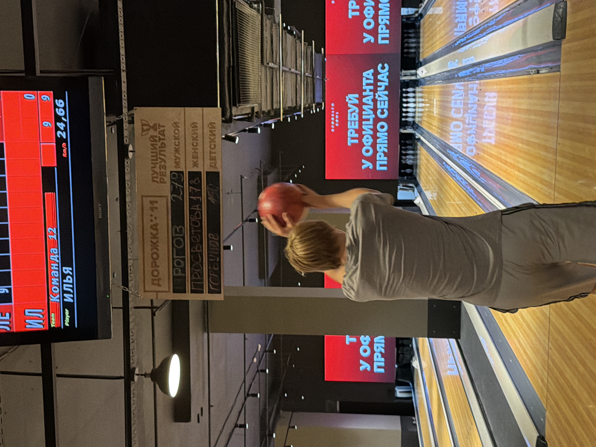 The image depicts a young man in athletic attire in the process of bowling. He is captured from behind, his right arm raised as he prepares to release a red bowling ball. In the foreground, bowling lanes are visible with pins set up at the far end.
In the background, a large screen displays a scoreboard. The scoreboard indicates a team name "ИЛЬЯ" with player "ИЛЬЯ", team name "Команда 12", and a score of "9". Another section of the scoreboard lists "ДОРОЖКА: 11" (Lane: 11) and "ЛУЧШИЙ РЕЗУЛЬТАТ" (Best Result). Below this, there are categories for "МУЖСКОЙ" (Men's), "ЖЕНСКИЙ" (Women's), and "ДЕТСКИЙ" (Children's) with associated numbers.
Further background details include overhead lighting fixtures and a sign that reads "ТРЕБУЙ У ОФИЦИАНТА ПРЯМО СЕЙЧАС" (Ask the waiter right now) on a large red screen. The overall environment suggests an indoor bowling alley. The text on the scoreboard and signs is in Russian.
The image does not contain enough information to determine the specific location within Izhevsk, Russia.