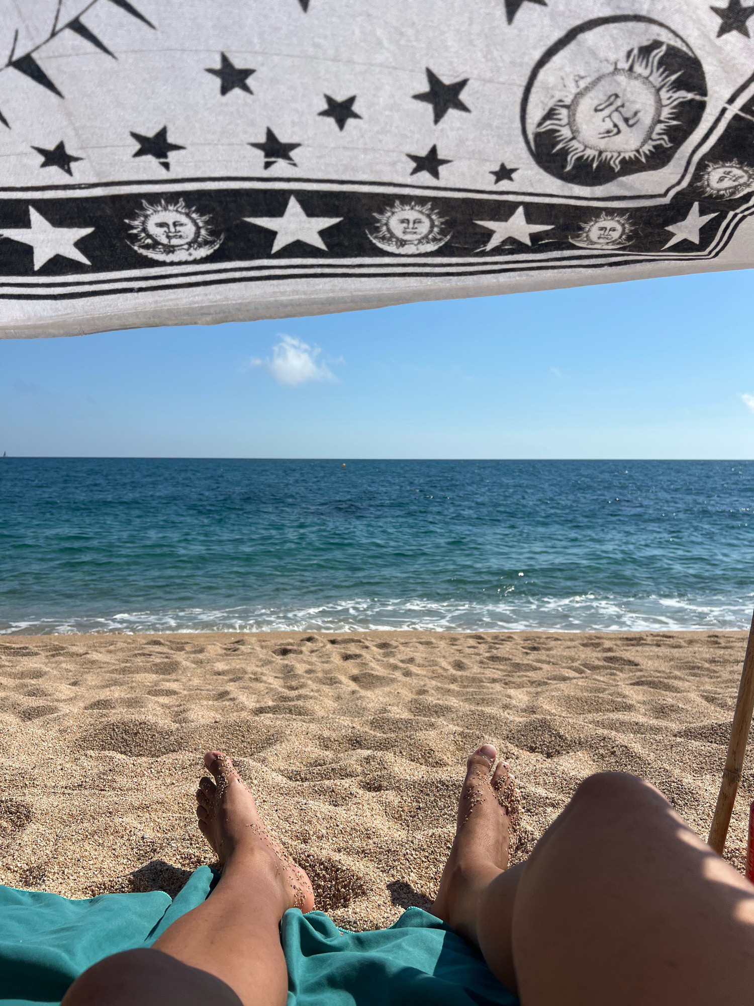 A first-person perspective shot from a beach blanket shows a person's legs and feet covered in sand, resting on a turquoise towel. In the foreground, the beach is covered in light brown sand. The ocean is visible in the midground with small waves and white foam rolling onto the shore. A clear blue sky with a few scattered white clouds is in the background. Overhead, a white canopy with a black pattern of stars and suns partially obscures the view of the sky. The scene is likely Sant Pol de Mar, Spain, given the characteristic Mediterranean blue water and sandy beach.