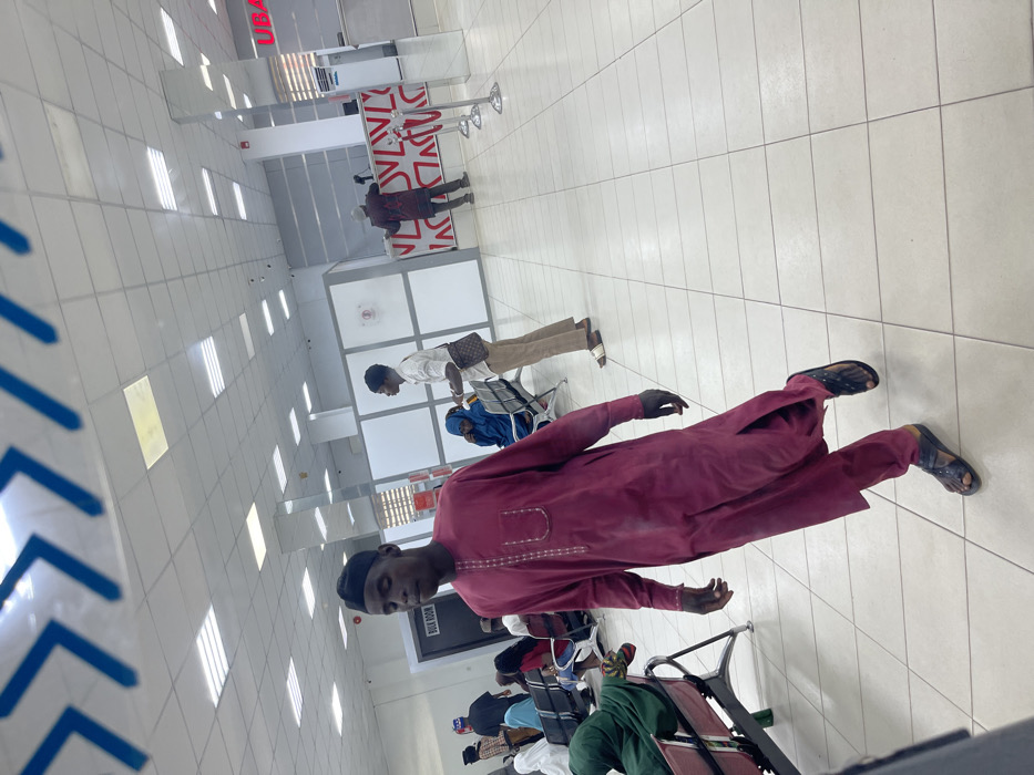 This image depicts the interior of a UBA bank branch in Zaria, Nigeria. In the foreground, a dark-skinned male wearing a maroon traditional tunic with embroidered patterns, matching trousers, and dark sandals is walking from left to right, his left foot mid-stride. His gaze is directed downwards and to his right.
In the midground, to the left, a row of metallic benches with red and green seating is partially visible, occupied by several individuals. A dark doorway marked "BULK ROOM" is present. To the right of the walking man, another dark-skinned male stands facing right, wearing a white long-sleeved collared shirt, light brown trousers, and light-colored sandals. He carries a dark-patterned crossbody bag on his right side. Behind him, a person in a blue head covering sits on a bench.
The background features bank counters with white and red patterned facades. A dark-skinned individual wearing a patterned shirt and dark trousers is standing at one of these counters, facing away from the camera, engaged in a transaction. Red stanchions with ropes are positioned in front of the counter. The "UBA" logo in red lettering is visible on a pillar or wall section to the far right. The environment is well-lit by numerous recessed rectangular fluorescent lights in a white tiled ceiling. The floor is also white-tiled with a grid pattern.
