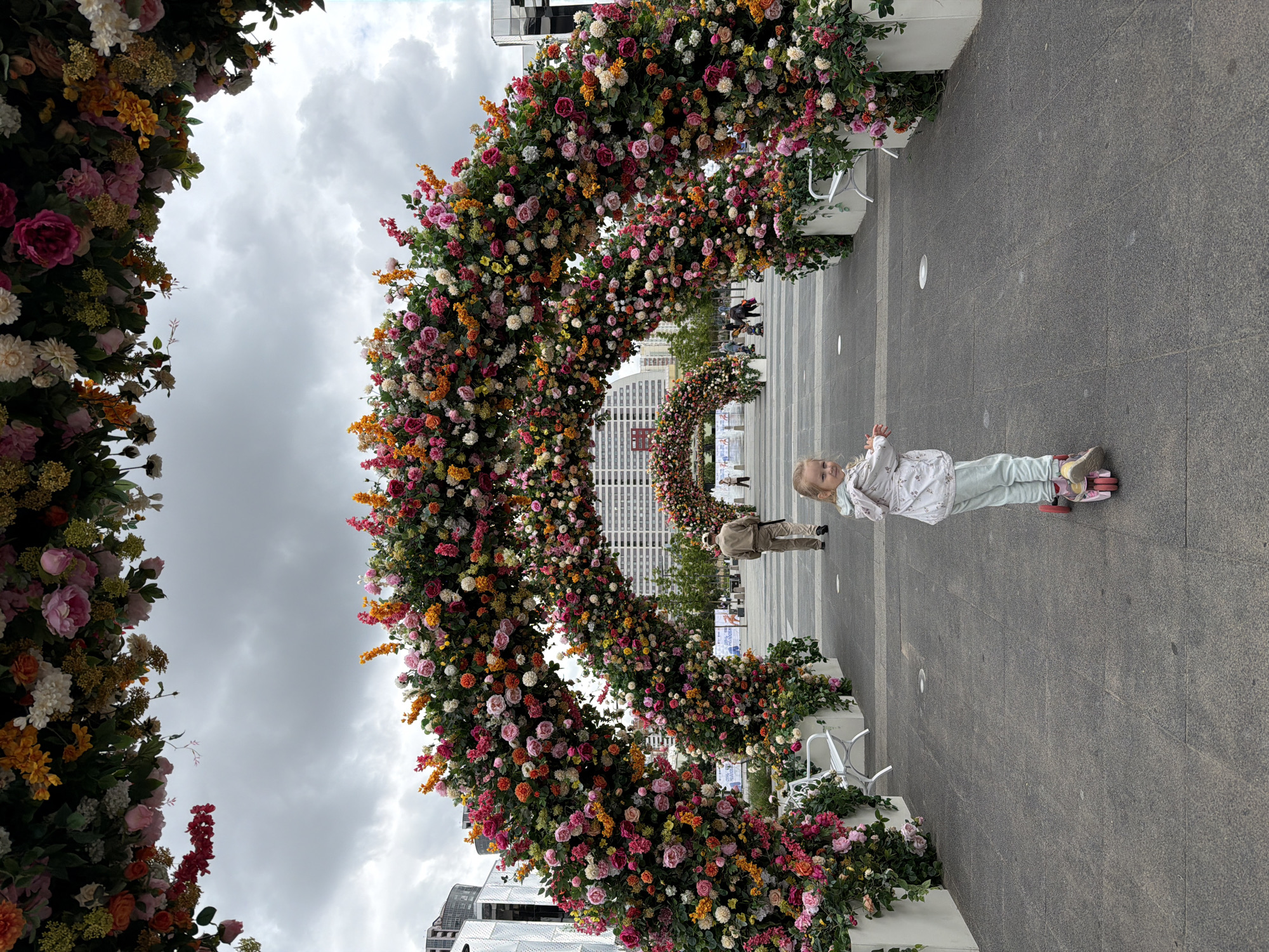 This is a vertical, full shot of an outdoor scene under a cloudy sky. The main subjects are a large, circular floral archway and a young child standing in front of it. The archway is densely packed with a variety of artificial flowers in shades of pink, orange, white, and yellow, with abundant green foliage. It extends across the width of the frame, creating a tunnel-like effect. Further down the pathway, a smaller, similar floral archway is visible.
A young child with blonde hair, wearing a light-colored patterned jacket and light blue pants, stands on the right side of the frame, looking towards the viewer and smiling. The child is positioned on a pink object, possibly a toy or a prop. Behind the child, a gray paved pathway stretches into the distance, lined with more floral arrangements and white planters. In the background, the facades of modern buildings with numerous windows are visible.
The scene appears to be a public space, possibly a park or plaza, decorated for an event or celebration. The cloudy sky suggests it is daytime, and the overall atmosphere is festive and cheerful due to the vibrant floral decorations and the child's presence. No visible text is present in the image. The location is identified as Khimki, Russia.