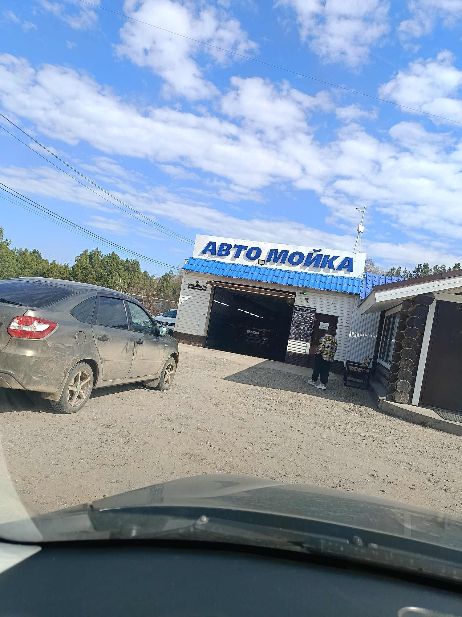 The image captures an outdoor scene in Chaikovsky, Russia, featuring a car wash facility under a bright blue sky dotted with scattered white clouds. The view is taken from inside a vehicle, with part of a dark dashboard and windshield visible in the immediate foreground.
In the midground, a dirty grey sedan, possibly a Datsun On-Do or Mi-Do, is parked on a dusty dirt and gravel lot, its rear facing slightly towards the viewer and its front towards the car wash entrance. Its tires and body are visibly covered in dirt, indicating a need for washing.
The main building is a single-story car wash with a blue corrugated roof and white walls. A large sign on the roof prominently displays "АВТО.МОЙКА" (CAR WASH) in blue Cyrillic letters. To the left of the car wash bay entrance, a detailed sign board is visible, which lists "АВТОШИНЫ, ШИНОМОНТАЖ" (CAR TIRES, TIRE FITTING) and a price list for various services. Inside the open car wash bay, a dark-colored car is partially visible.
A person dressed in a plaid shirt, dark pants, and light-colored shoes stands near the car wash entrance, facing the service area, possibly waiting for service or a vehicle.
To the right of the car wash structure, there is an adjacent rustic-style building made of logs, featuring a brown roof and a door with a sign that reads "ОТКРЫТО" (OPEN). This building appears to be an office or waiting area for the car wash.
In the background, a line of green coniferous trees stretches across the horizon, and several power lines are visible against the clear sky. The ground is dry and unpaved. The scene depicts a typical bright, sunny day at a roadside service establishment.