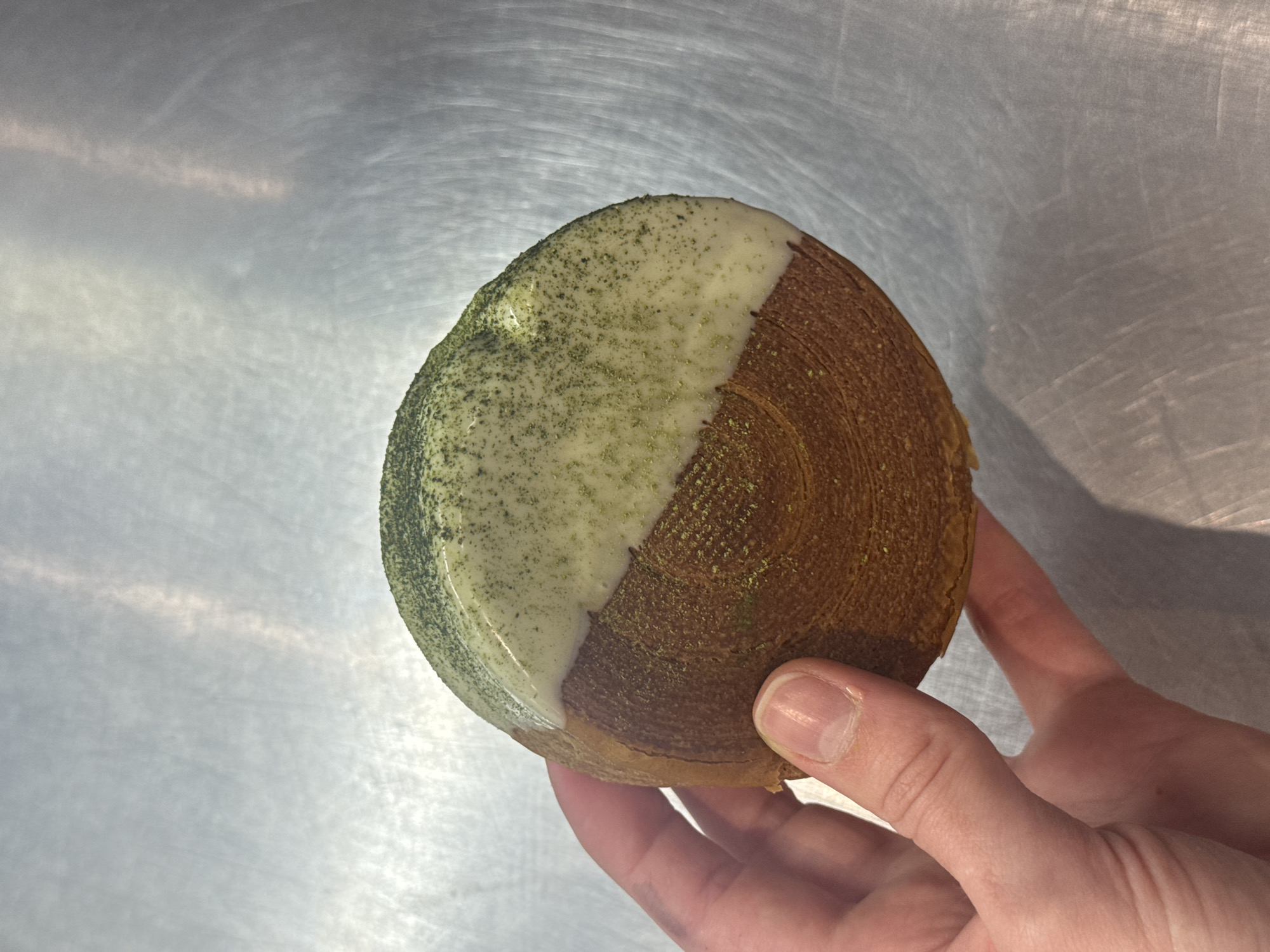 A hand holds a round pastry, likely a croissant or similar baked good with visible layers. Half of the pastry is coated in a white glaze, which is speckled with green powder, potentially matcha. The other half is golden brown with visible concentric rings of layers. The background is a brushed metal surface. The image does not contain any information about its location in Vracar Urban Municipality, Serbia.