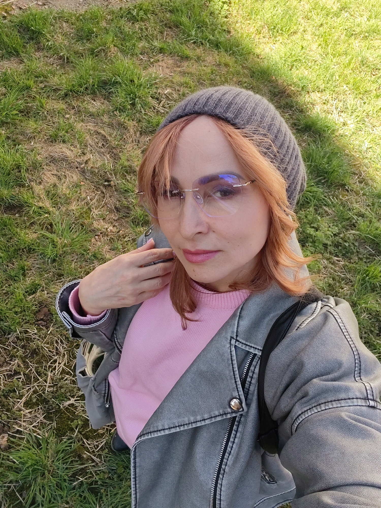 A person is taking a selfie outdoors. The background shows grass, suggesting a park or green space. The person is wearing a gray beanie, eyeglasses, a pink sweater, and a gray leather jacket. A strap, possibly from a bag, is visible over the shoulder. The location, Chaikovsky, Russia, is not discernible from the image alone.