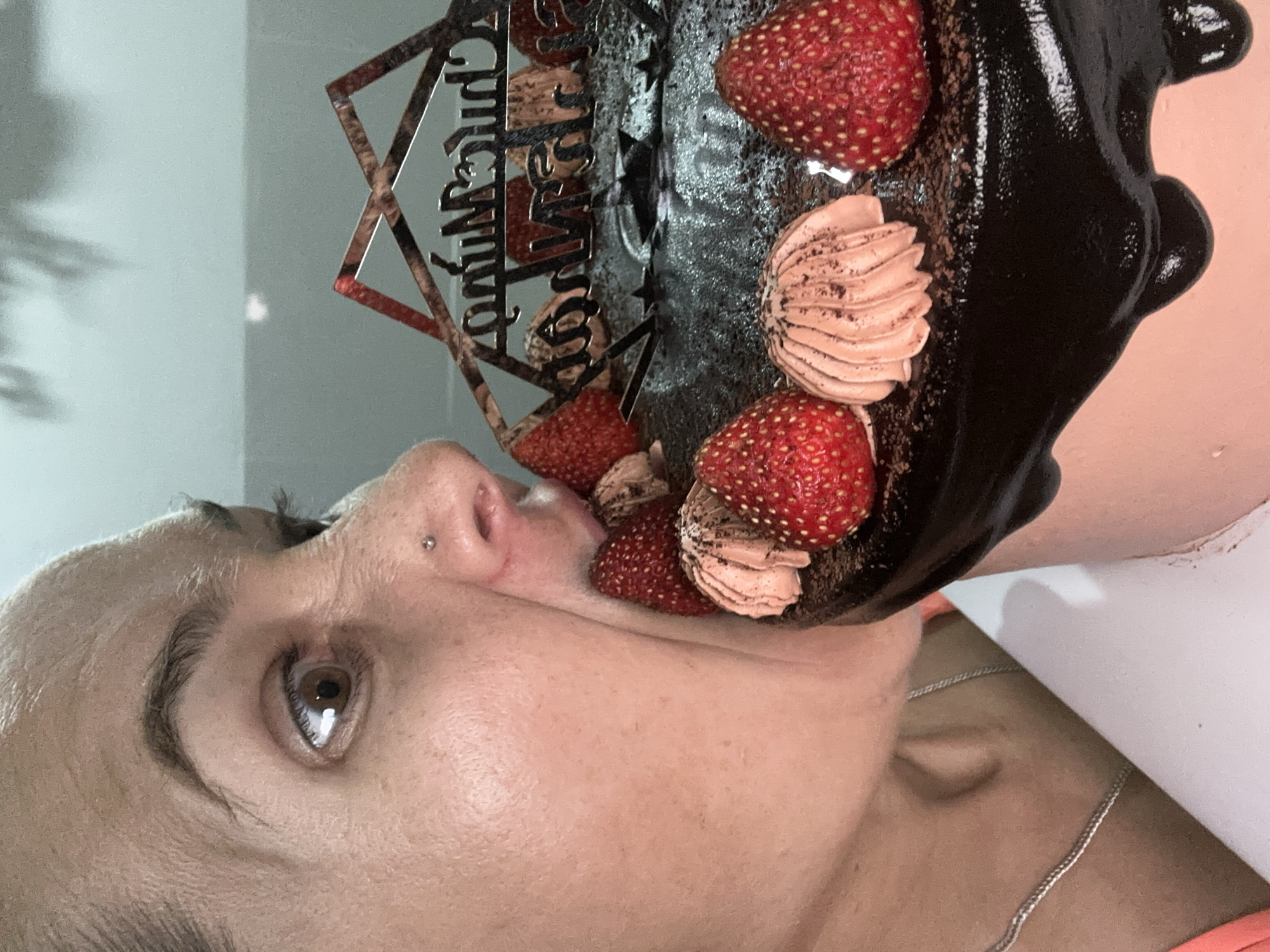 A woman is shown in a close-up, portrait-style view, holding a chocolate cake close to her face. The cake is decorated with strawberries and frosting swirls. A cake topper with text is visible. The setting suggests an indoor location. The image provides no specific information to place the scene in Bac Nha Trang Ward, Vietnam.